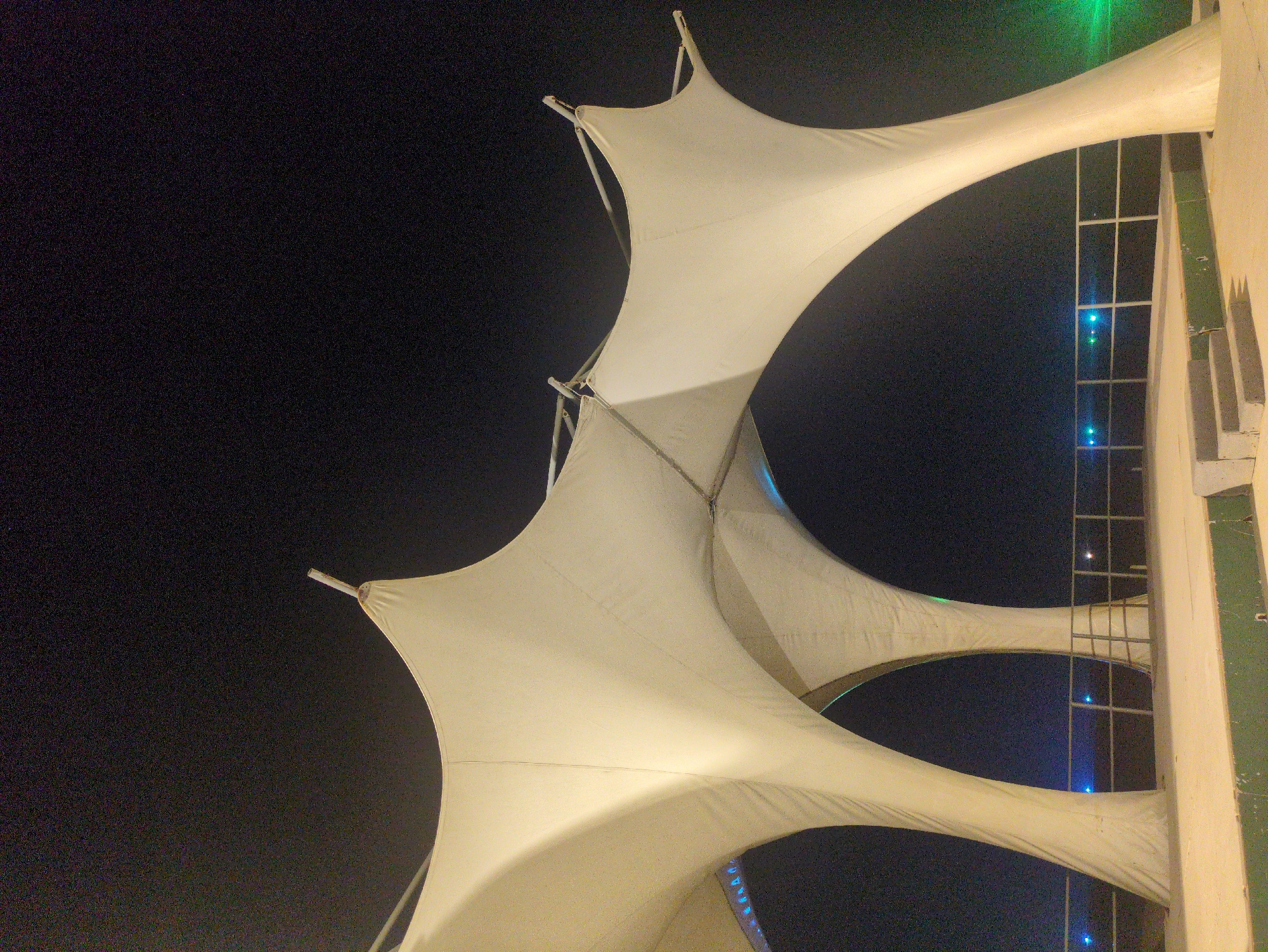 Captured at night in Phú Quốc, Vietnam, the image presents a low-angle view of a multi-layered, white fabric structure. The fabric is taut, stretched between support points, creating a dynamic, almost organic shape against the dark sky. The fabric structure is situated adjacent to a building with a grid-like window design, illuminated with blue lights. A green light source is visible near the building. The structure appears to be architectural.