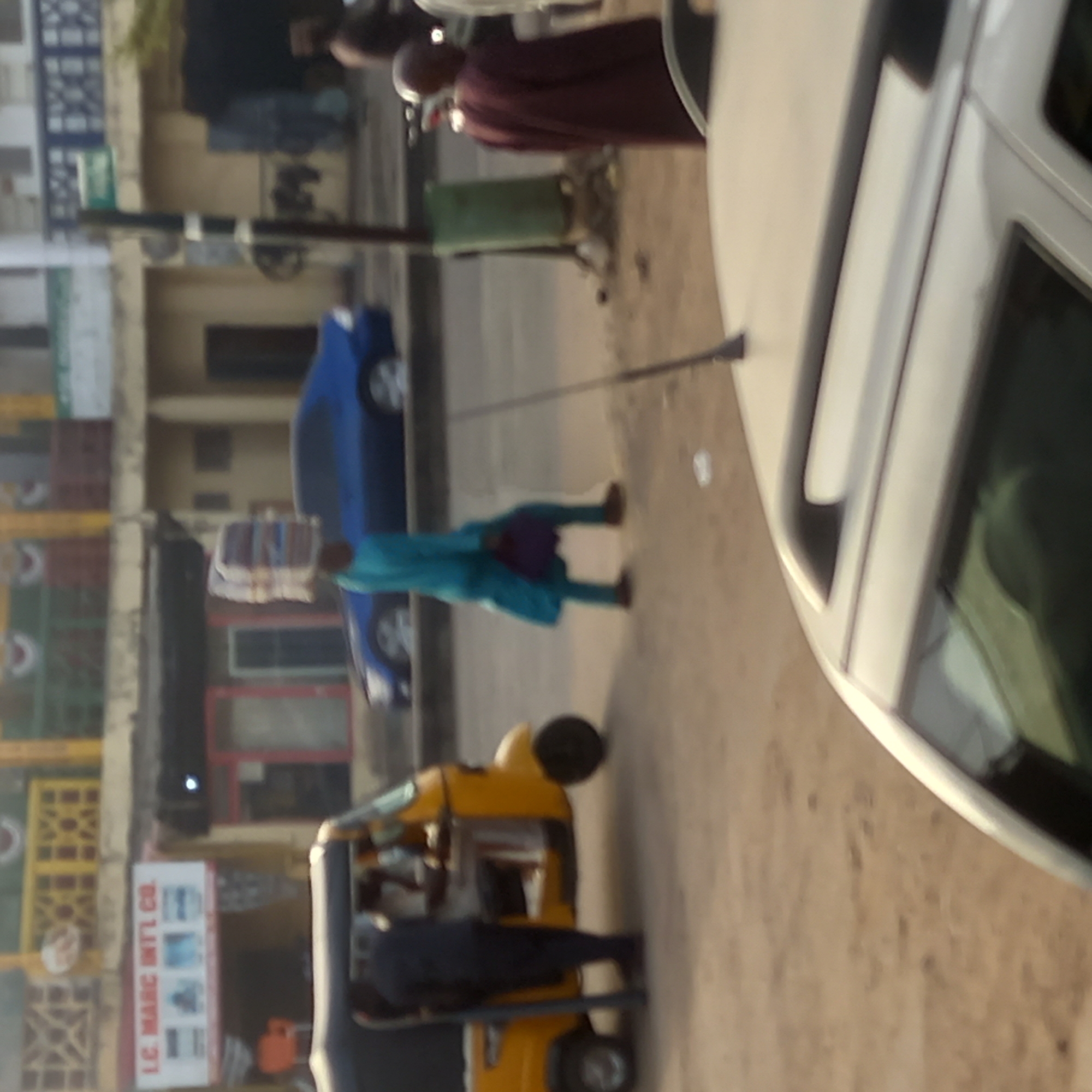 The image shows a street scene in Dadi Riba, Nigeria. A yellow auto-rickshaw (keke) is parked on the side of the street, with people inside. A man in a blue robe walks past a parked blue car, carrying a load on his head. Another man in a brown robe walks along the street further ahead. A portion of a white car is visible in the foreground. Buildings with various shops and signboards line the street.