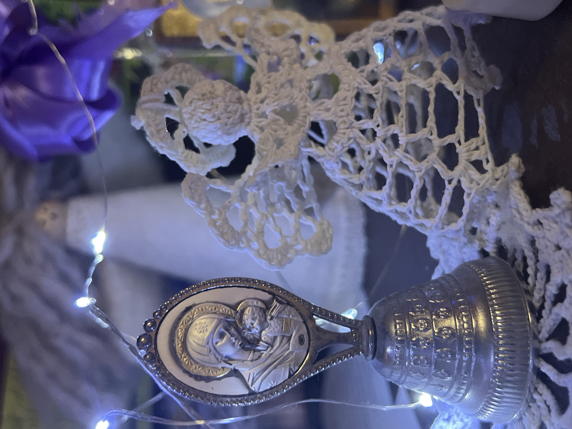 The image shows a close-up of Christmas decorations. In the foreground, on the left, is a silver bell with an oval-shaped medallion on top, depicting an image of the Virgin Mary holding the infant Jesus. To the right of the bell is a white crocheted angel figure. The angel has its arms raised and its wings spread. The decorations are illuminated by a strand of small, white LED lights, which are visible woven throughout the scene. In the background, out of focus, are other decorations, including what appears to be a larger, cone-shaped angel or Christmas tree topper, and a swirl of purple fabric. The overall lighting is dim, creating a festive and slightly ethereal atmosphere. The scene does not provide enough information to determine its location in the city of Minsk, Belarus.