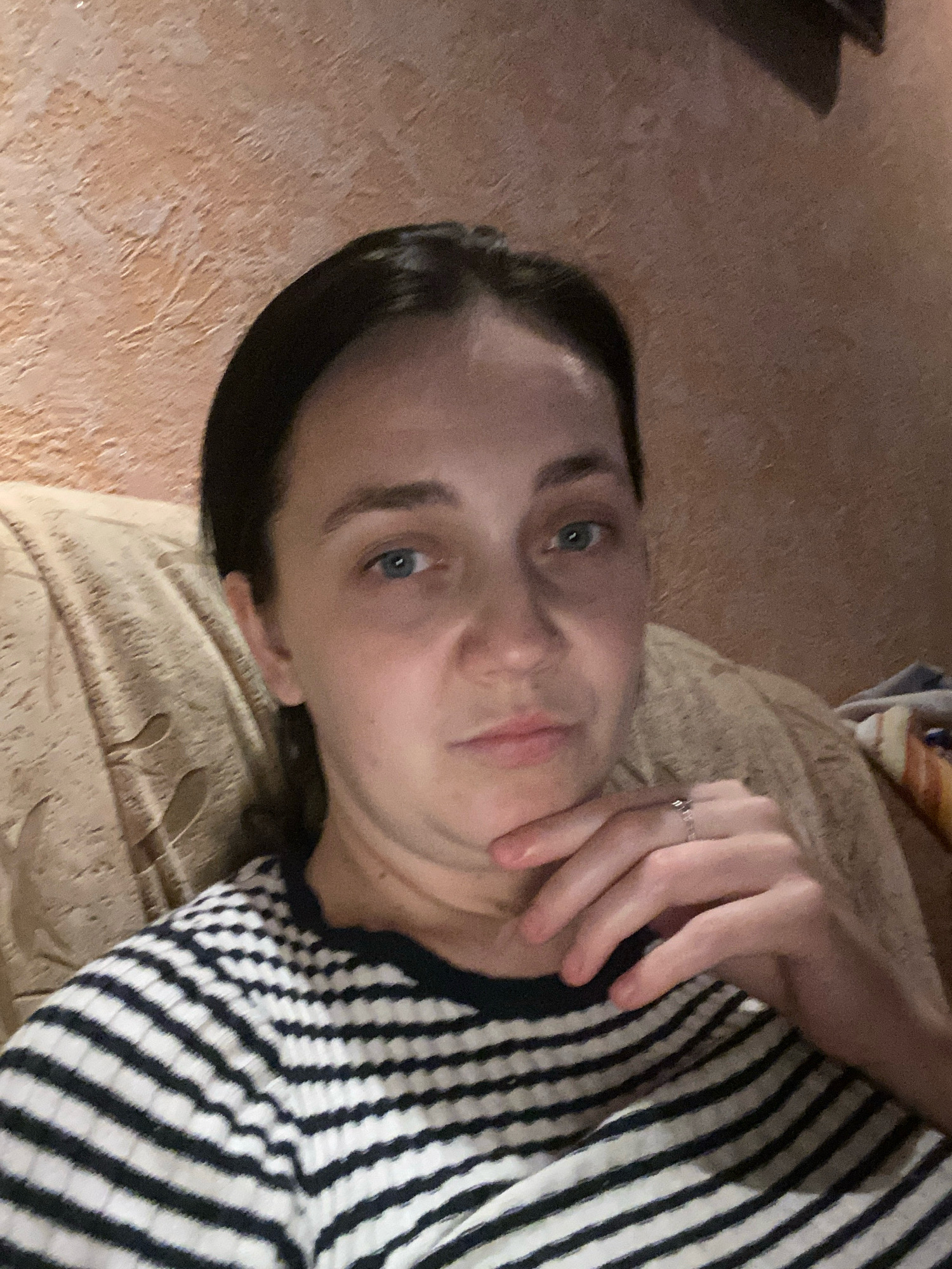A medium shot shows a woman with dark hair, wearing a black and white striped shirt and a ring on her right hand, sitting on a couch with her chin resting on her hand. The background consists of a textured, peach-colored wall and a portion of a patterned couch. The lighting is dim. The image appears to be a selfie.