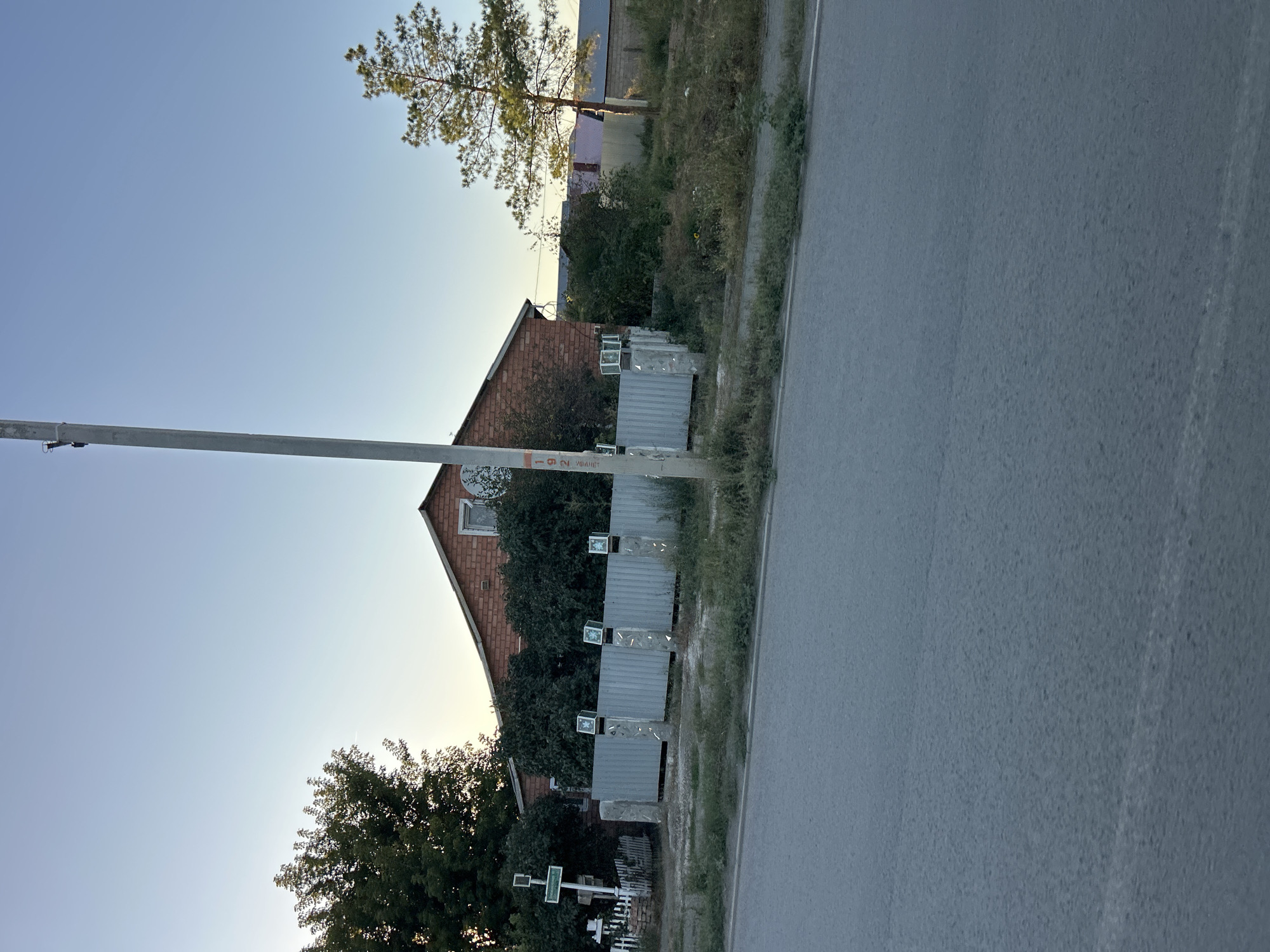A slightly angled medium shot captures a scene in Orsk, Russia, under a clear blue sky during daylight hours. In the foreground, a paved road with a white line marking its edge occupies the bottom third of the frame. Beyond the road, a strip of grass and weeds separates it from a metal fence. The fence, composed of vertical corrugated metal panels, is supported by stone pillars. Mounted on some of these pillars are small, square glass boxes with what appear to be images or lights inside.
Behind the fence, a low-rise brick building with a gabled roof is visible. A window is present in the upper part of the gable. Dense green foliage partially obscures the building and the area behind it. To the left of the building, a tall, slender pole stands vertically. Attached to the pole is a white sign with green lettering. Further left, a white picket fence encloses a small area. A tall tree with dark green foliage is situated to the left of the building, and a thinner pine tree stands to the right. In the background, more buildings with colorful roofs are partially visible. The overall impression is of a residential area with established trees and fencing.