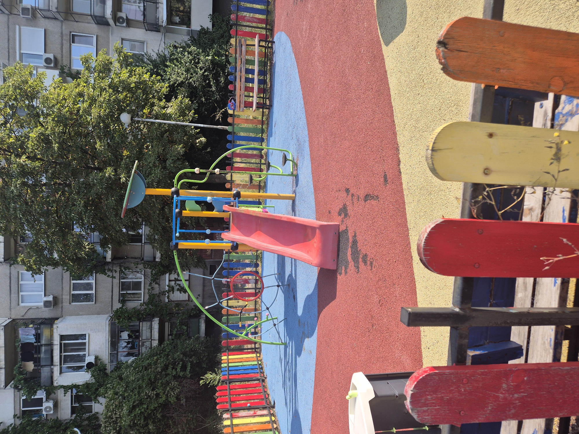 The image depicts a children's playground situated in front of a Soviet-era apartment building, typical of neighborhoods in Sofia, Bulgaria. The playground features a red slide, a climbing frame with green and yellow poles, and a rainbow-colored fence. The ground is a mosaic of red and blue safety surfacing. A wooden bench is visible behind the fence. The apartment building is grey concrete with multiple balconies, some with clothes drying on them, and appears somewhat dilapidated. Greenery from trees and vines partially obscures the building, suggesting an urban setting with established vegetation. The scene is bathed in bright sunlight, casting shadows on the playground. The perspective is from ground level, looking towards the playground and the building.