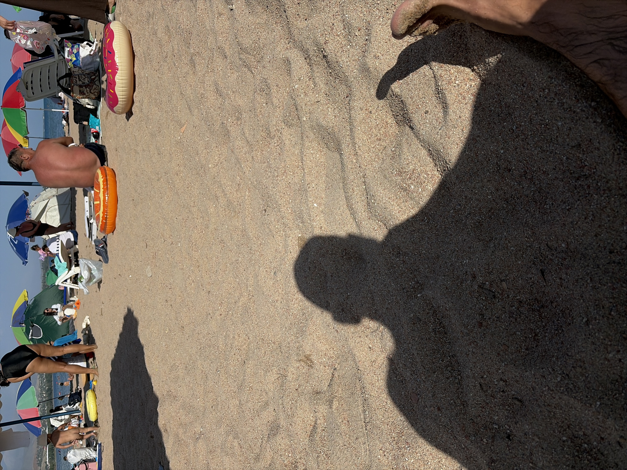 The image depicts a sandy beach scene in Cholpon-Ata, Kyrgyzstan. In the foreground, the shadow of a person's head and torso is cast on the sand, with a bare foot also visible in the lower right corner, covered in sand. Further back on the beach, several people are present. A woman in a black swimsuit stands near the water, facing away from the camera. To her left, a young child, also nude, stands on the sand. Other beachgoers are seated or lounging under umbrellas in the background, with beach chairs and inflatable rings scattered on the sand. The environment suggests a sunny day at a resort area, with beach umbrellas, tents, and lounge chairs indicating recreational use.