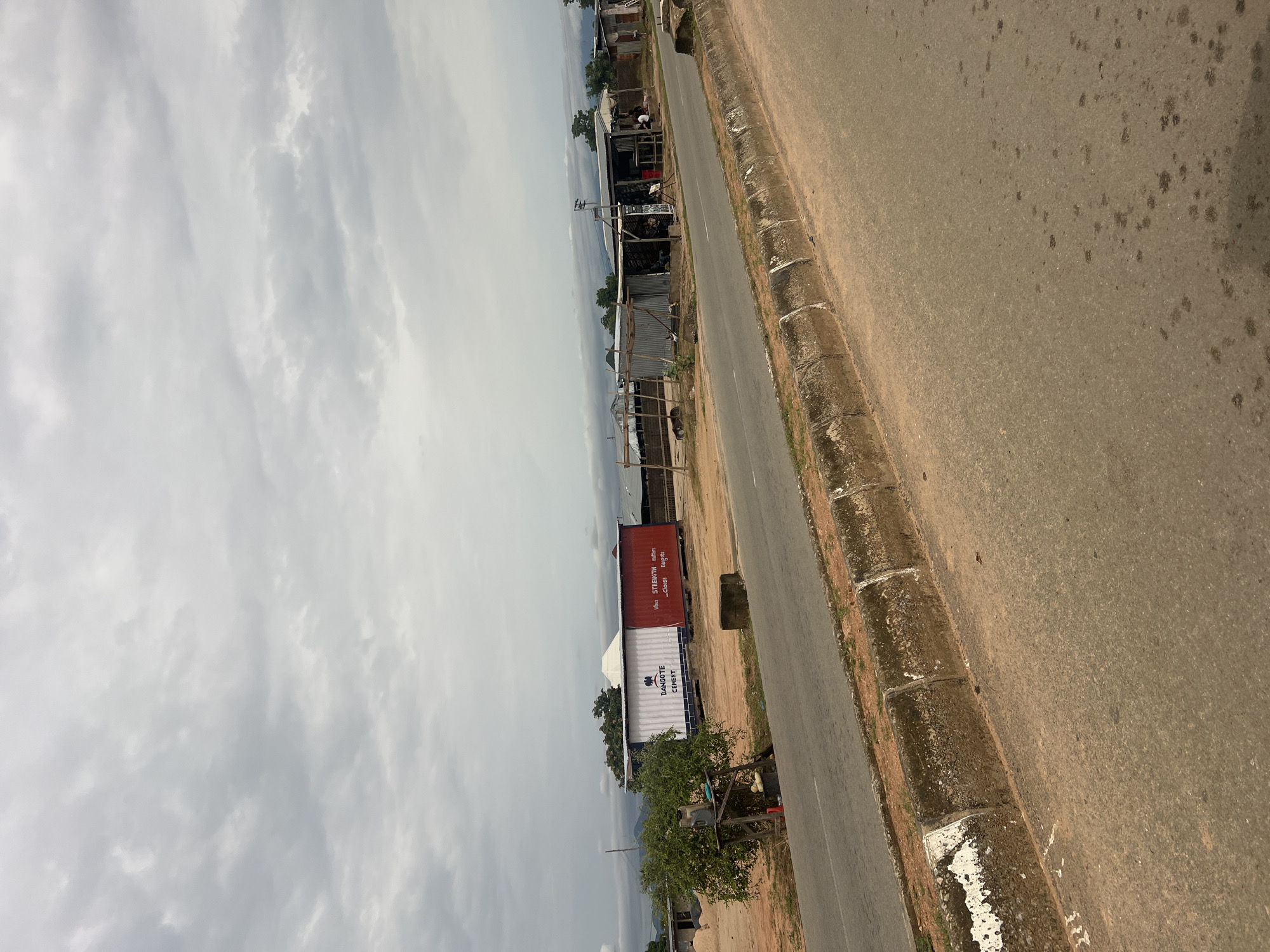 The image shows a rural roadside scene in Jalingo, Nigeria, under an overcast sky. A paved road runs diagonally through the frame, bordered by a rough, dirt shoulder with some vegetation and scattered debris. Along the left side of the road, there are several small, open-air structures that appear to be shops or market stalls, constructed with corrugated metal and wooden frames. Some of these stalls have goods displayed outside, and a few people are visible, seemingly engaged in daily activities, though their specific actions are unclear.
Dominating the mid-ground are two large shipping containers. The top container is red and has white text that is difficult to read due to the angle and resolution. Below it is a white container with blue lettering that clearly reads "DANGOTE CEMENT." A small tree grows near the containers. Further down the road, more structures and trees are visible, suggesting a scattered settlement. The overall impression is one of a functional, somewhat weathered, rural commercial area on a cloudy day. The time of day appears to be daytime, but the cloudy conditions obscure direct sunlight and create a diffused light.