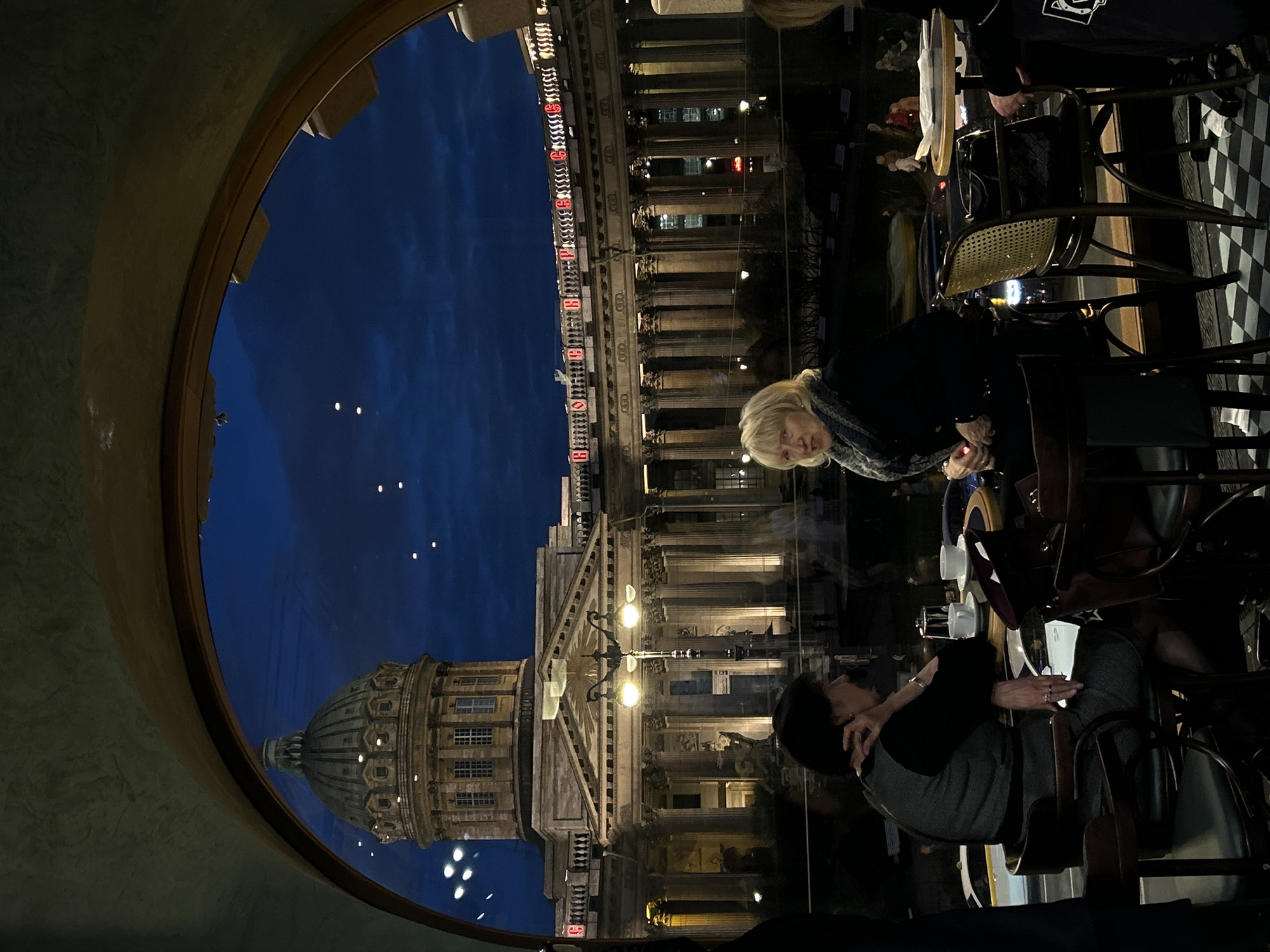 The image portrays a nighttime scene viewed from inside a cafe or restaurant in Saint Petersburg, Russia. The perspective is through a large, circular window, framing a view of illuminated city architecture under a deep blue evening sky.
Inside, two women are seated at tables. In the foreground, a blonde-haired woman, wearing a patterned scarf, is visible from the chest up, looking towards the window. Further back, another woman with dark hair is seated, also looking outwards or at a companion. The interior is dimly lit, featuring dark, textured walls and classic cafe furniture, including wooden chairs with wicker backs.
Outside the window, a prominent cityscape unfolds. On the left, the distinctive dome of Kazan Cathedral is visible, its structure illuminated against the darkening sky. Directly across the street, a grand, multi-storied building with numerous classical columns and a pediment stands brightly lit. On its facade, illuminated in red, the word "CHOCOLATE" is clearly legible, likely advertising a business, with the number "8" also visible. Streetlights and other distant building lights twinkle in the scene. The overall ambiance is tranquil and atmospheric, capturing an evening moment in a historic city.