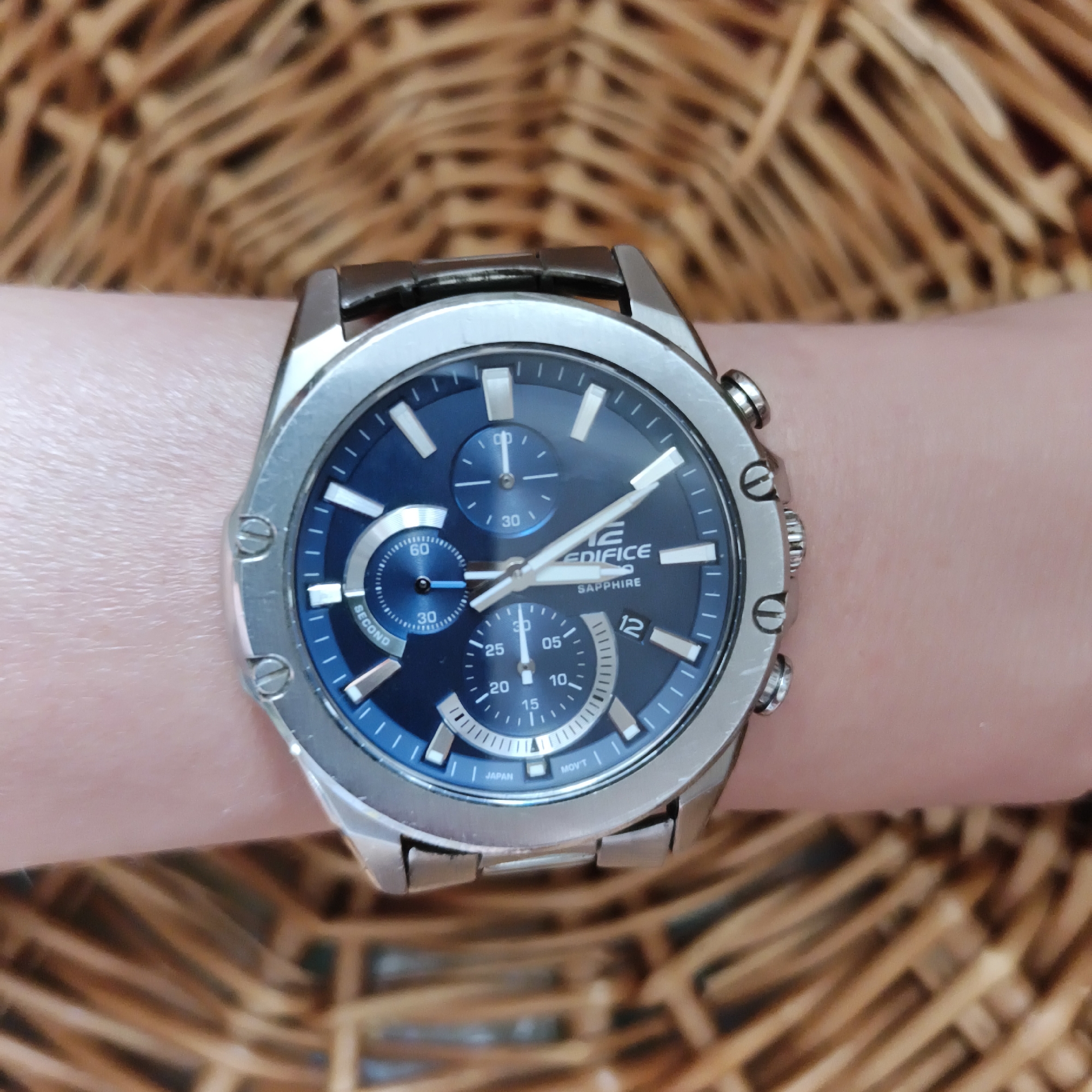 A man's wrist, adorned with a silver-toned Edifice watch with a blue dial, is positioned over a woven basket. The watch features multiple subdials, including one indicating seconds with the inscription "SECOND" and another that appears to be a chronograph. The watch face also displays the text "EDIFICE SAPPHIRE" and "JAPAN MOV'T". The basket's pattern is clearly visible, creating a textured backdrop. The image is a close-up, focusing on the watch and wrist, with the background being slightly out of focus. There is no discernible location within the city of Chaikovsky, Russia.