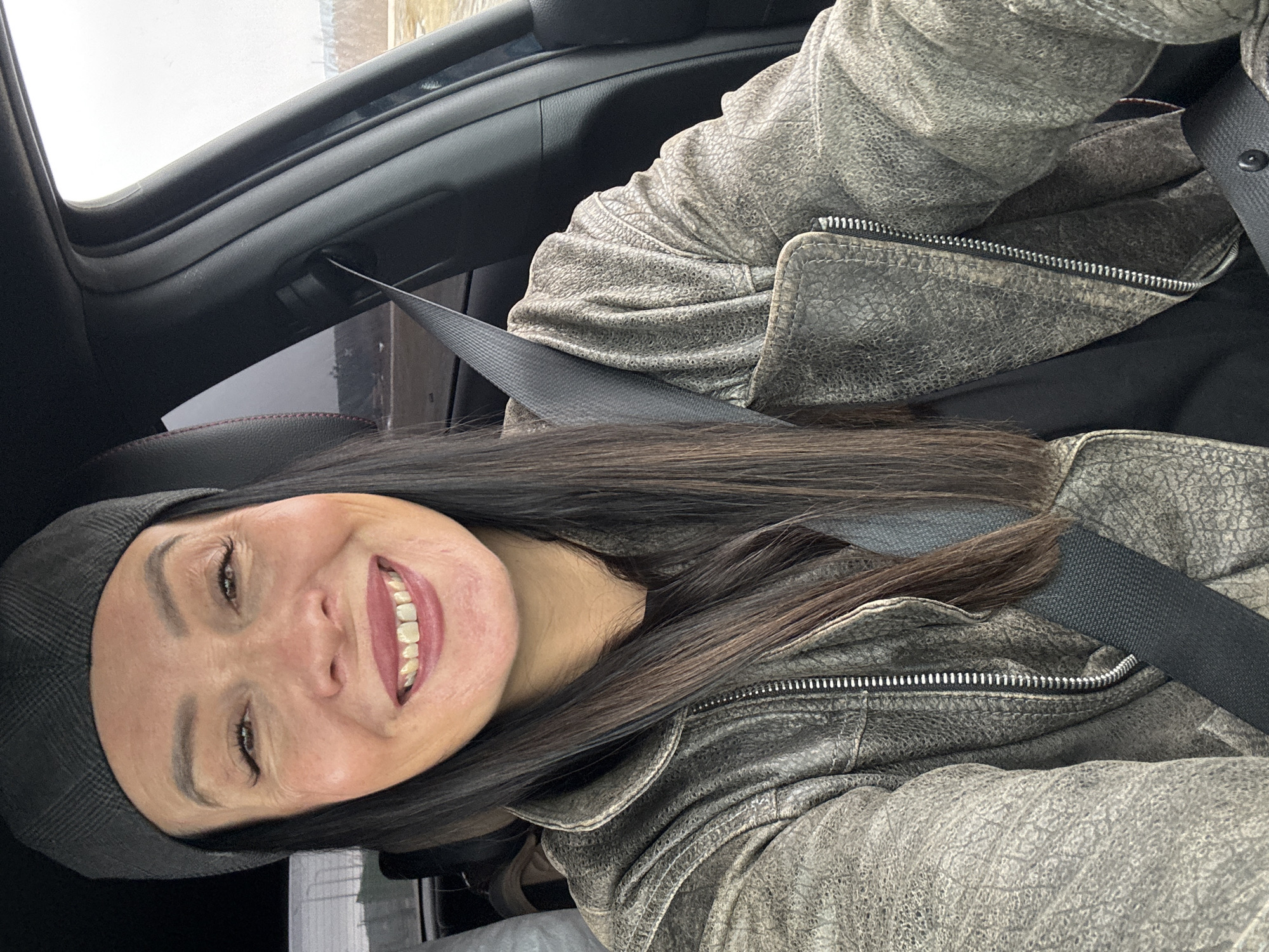 A woman is inside a car. She is wearing a grey leather jacket and a dark plaid beret. She is smiling and wearing a seatbelt. The background is out of focus, but appears to be a street scene. The image was not taken in Chaikovsky, Russia.