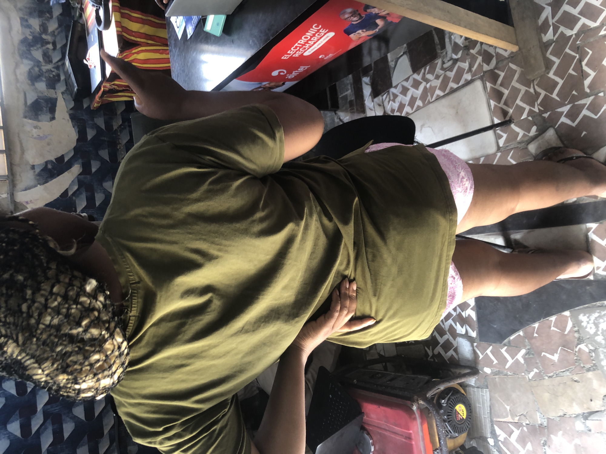 A medium shot captures a woman from the rear, standing in what appears to be a business establishment in Ado, Nigeria. She is wearing a green t-shirt and pink patterned shorts. Her right hand is placed on her lower back, and her left hand is raised, gesturing towards something out of frame. Her hair is covered with a dark net. In the background, a counter is visible with a laptop, headphones, and various papers. A red and yellow striped fabric is draped over a table behind the counter. The floor is tiled with a brown and white pattern. To the left of the woman, a red and black generator is present. An "Airtel ELECTRONIC RECHARGE" sign is affixed to the counter.