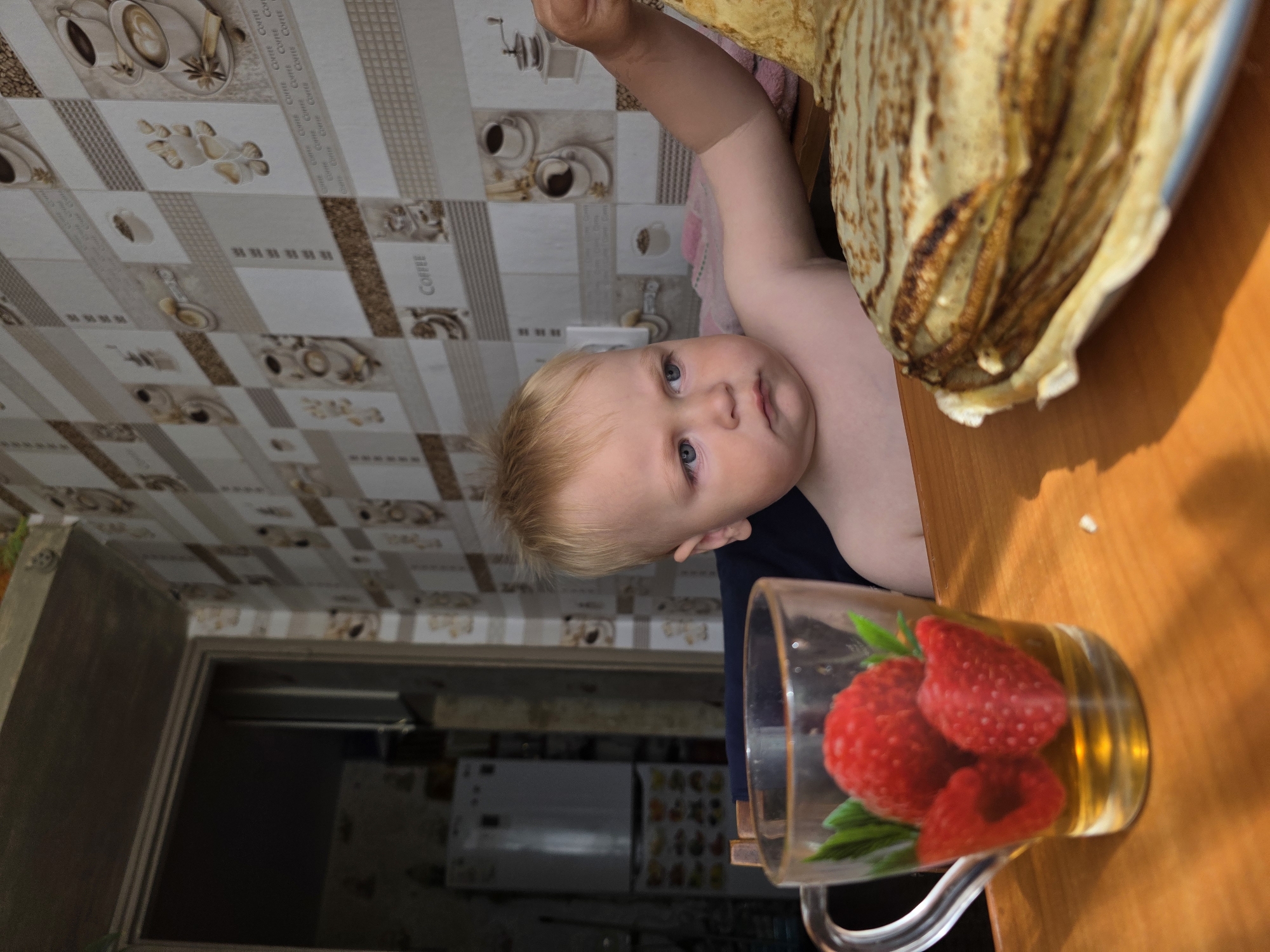 A young Caucasian toddler with blonde hair and blue eyes sits at a wooden table in a room with coffee-themed wallpaper. The toddler is looking to the right with a slightly furrowed brow. In the foreground, a glass mug with a handle contains a golden liquid with three decorative raspberries and green leaves. To the right of the mug is a large stack of thin, golden-brown pancakes on a plate. A white refrigerator is visible in the background through an open doorway. The overall setting appears to be a domestic kitchen in Chaikovsky, Russia.