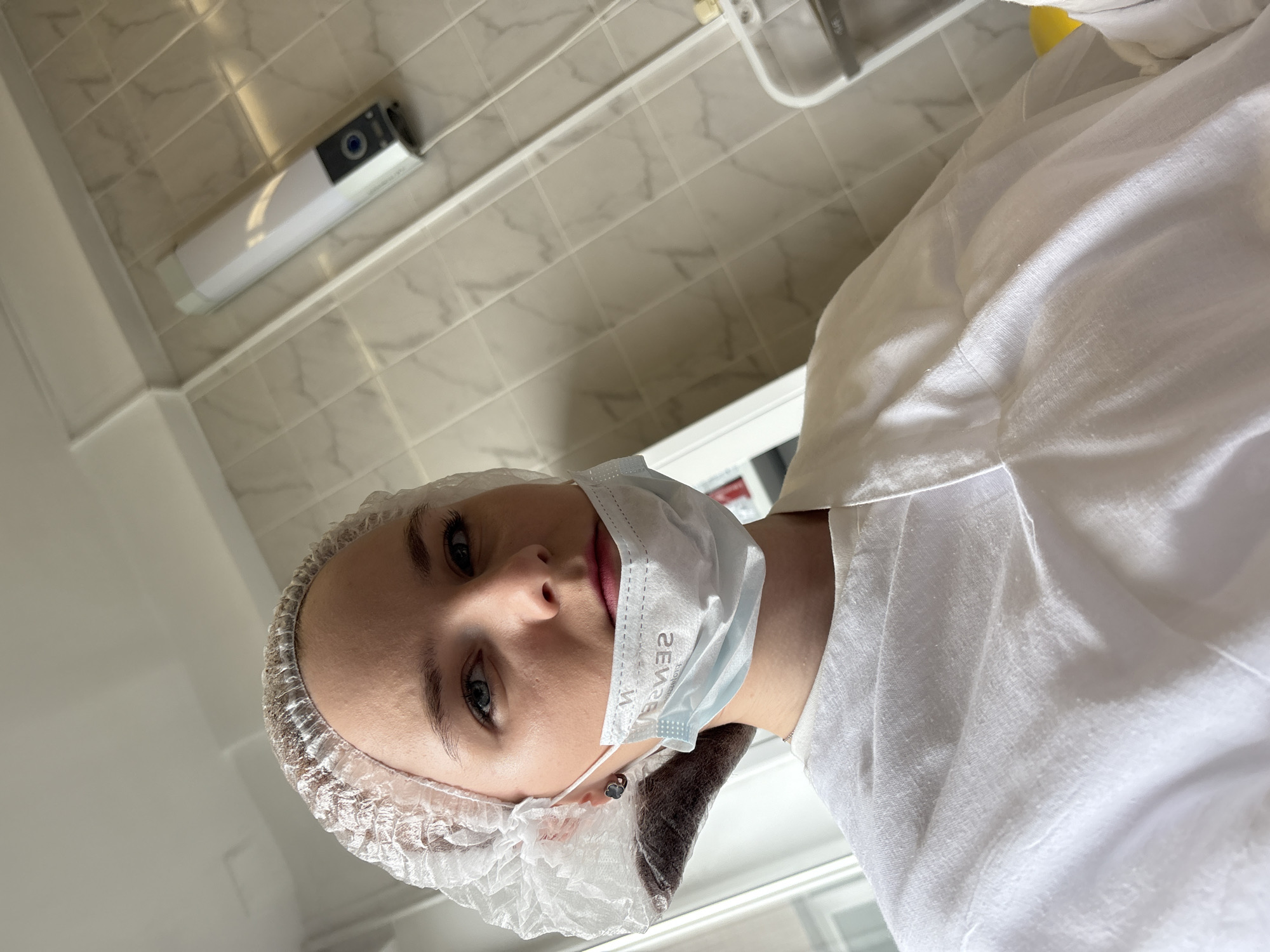 A portrait-style photograph presents a person in what appears to be a healthcare setting. The individual is wearing a white bouffant cap and a light blue surgical mask. A white garment covers their upper body. The background reveals a tiled wall with a rectangular object mounted on it. The location is indicated as Saint Petersburg, Russia.