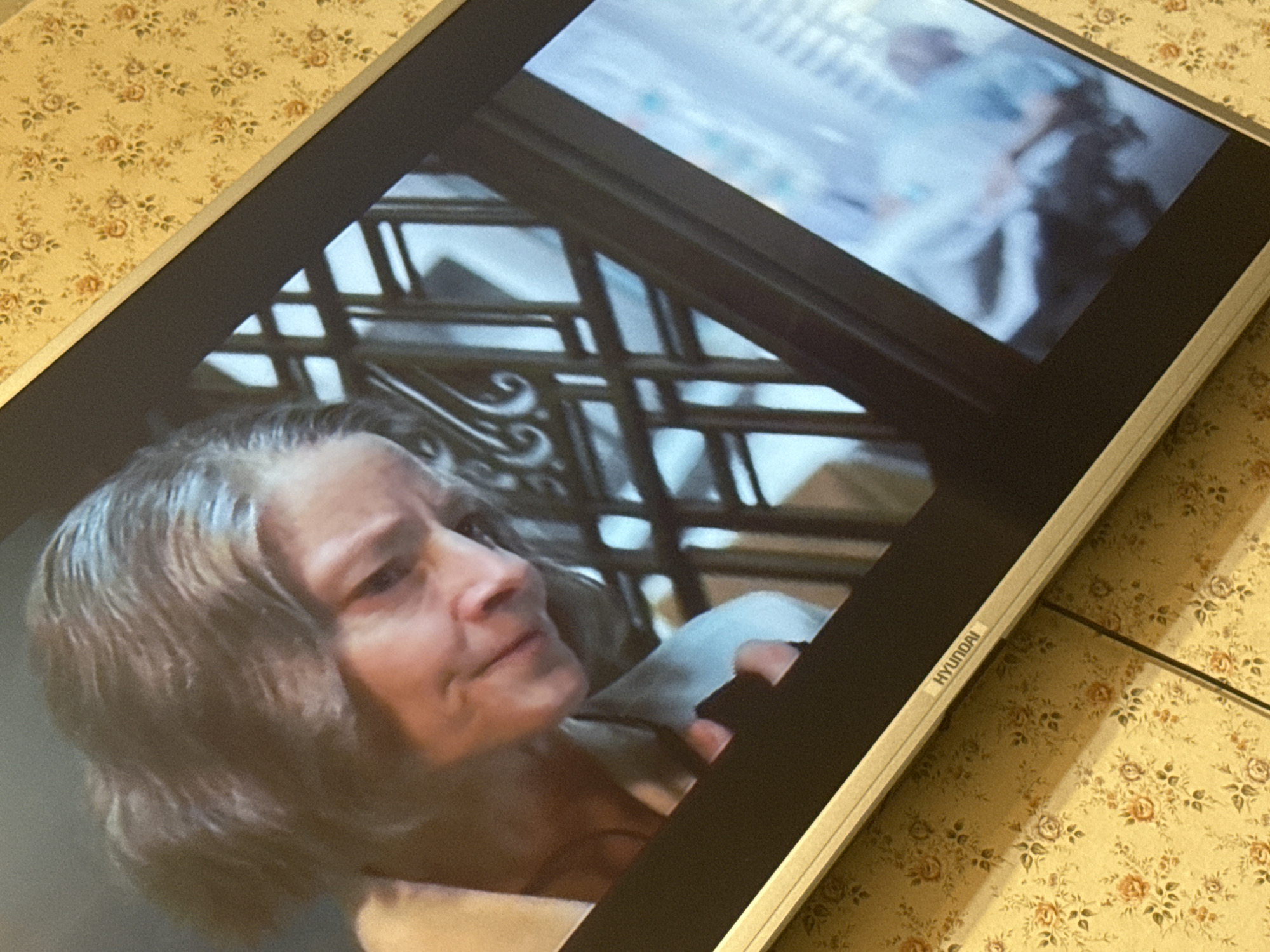 A Hyundai monitor displays a scene depicting a woman with graying hair, presumably indoors near an elaborate wrought-iron staircase. The monitor rests on a surface covered with a light yellow floral patterned material. The scene's location cannot be determined to be in Pushkino, Russia without further evidence.