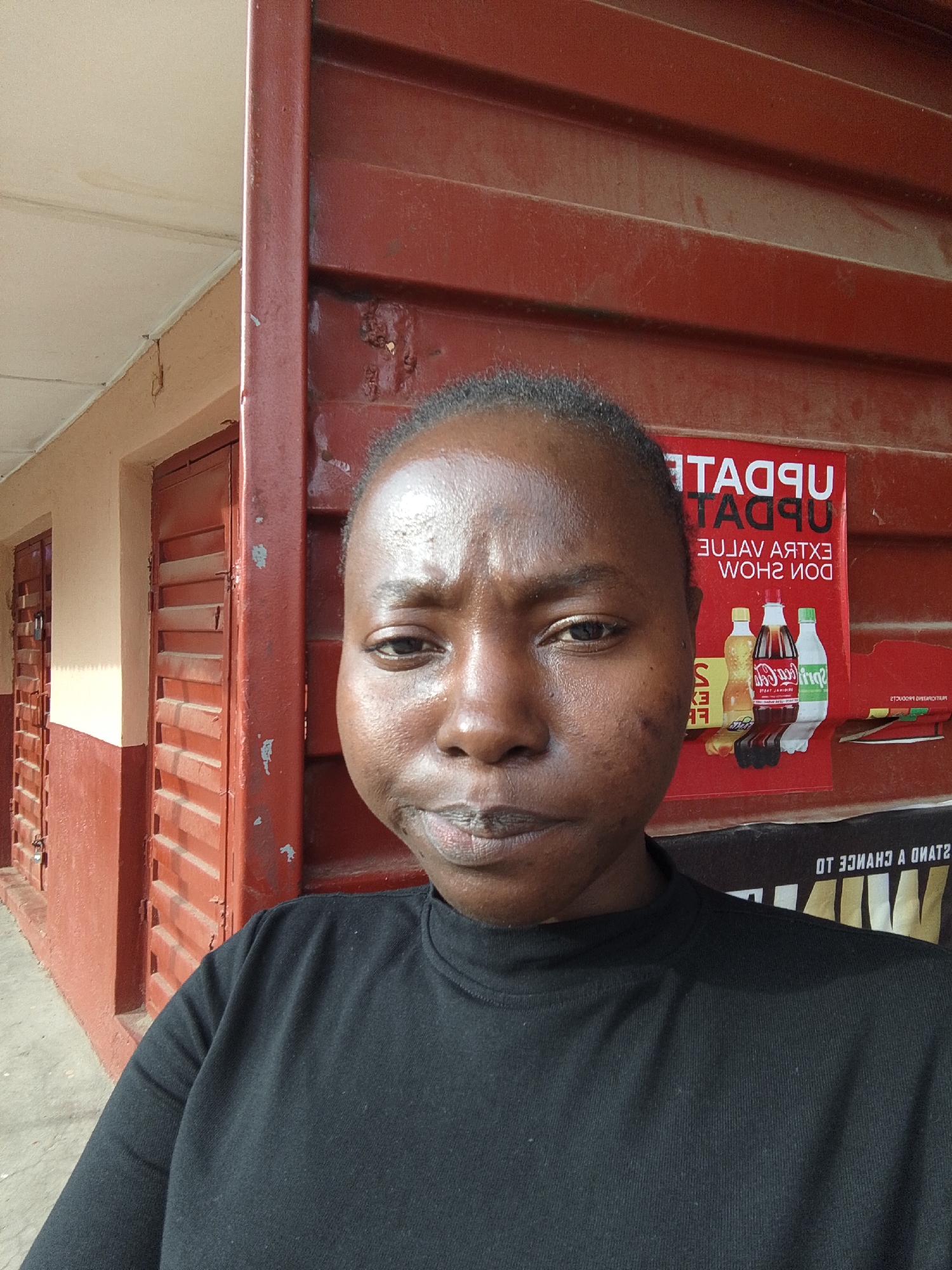 This is a medium shot, selfie-style photograph of a young Black woman taken outdoors. She is facing the camera with a neutral expression, her head slightly tilted to her left. She is wearing a black, long-sleeved, high-neck shirt. The background features a red corrugated metal wall, with a poster that has text in red and white, and images of beverage bottles. The poster appears to advertise a "Value Extra Value Don Show" and displays Coca-Cola and Sprite bottles. To the left of the woman, there are two doors with slatted fronts, painted in red and a lighter beige/tan color. The overall setting appears to be an outdoor space, possibly in front of a building or shop. The lighting suggests it is daytime, with sunlight casting shadows. Visible text includes "UPDATE" in large letters on the poster, and other fragmented text in what appears to be English, though some is reversed or partially obscured. The woman's skin is dark and appears slightly shiny, possibly due to sweat or lotion. Her hair is pulled back, and her eyebrows are dark. There are some small dark marks on her cheeks.