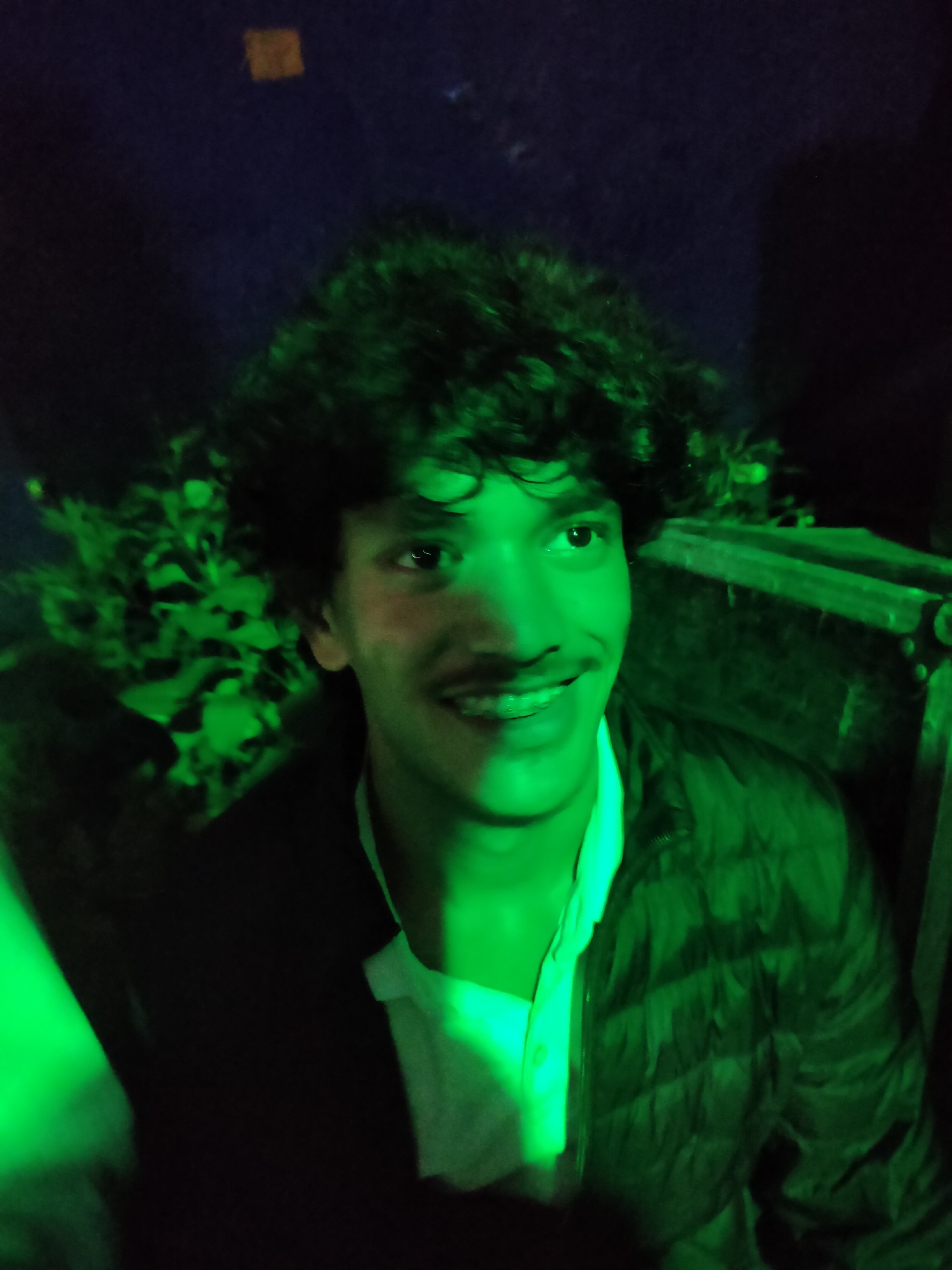 A young man with curly dark hair and a mustache smiles, revealing braces. He is wearing a white collared shirt under a dark, puffy jacket. The lighting is predominantly green, casting an eerie glow on his face and the surrounding environment, which includes what appears to be foliage and a dark, textured surface in the background. The setting is dimly lit, with the green light dominating the scene. There is no discernible indication of the city of Damadi, India, within the image.