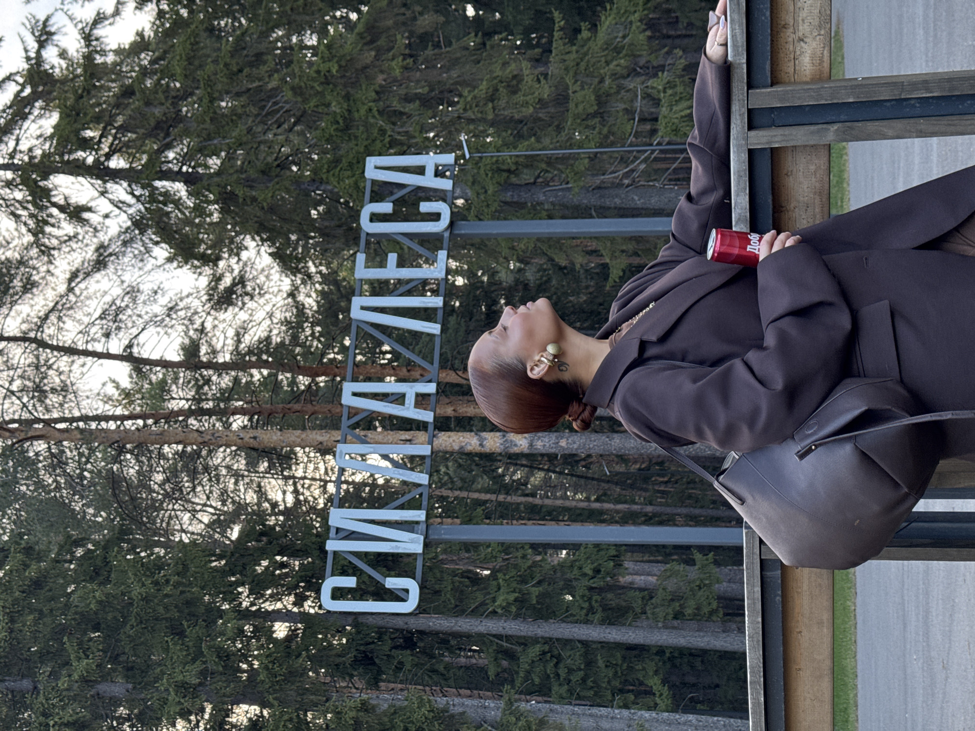 A woman in a brown blazer holds a red can of soda and poses near a sign that reads "СИЛА ЛЕСА," which translates to "The Power of the Forest." The sign is positioned in front of a backdrop of tall trees. This is likely located in Votkinsk, Russia.