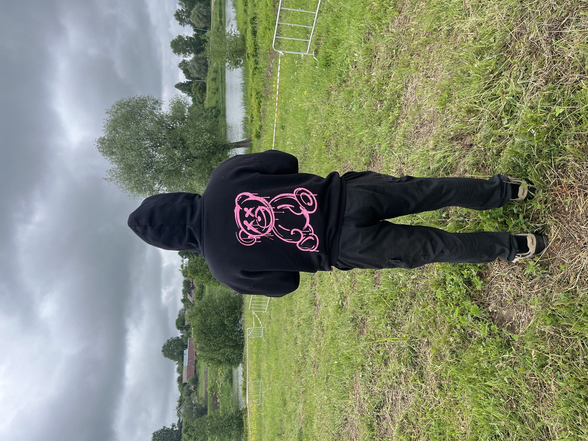 The image captures an individual standing with their back to the camera, positioned outdoors in a rural or semi-rural setting. The person is wearing a black hooded sweatshirt with the hood up; the back of the sweatshirt features a distinctive bright pink graphic of a teddy bear with 'X's for eyes and a distressed, dripping aesthetic. They are also wearing black cargo-style pants and beige and black athletic shoes, with a brand logo visible on the left shoe. The individual stands on uneven terrain covered in green and dry grass, with small patches of wildflowers.
In the mid-ground, a temporary barrier is present, constructed from white metal crowd control fences linked by white and red striped tape, extending horizontally across the scene. Beyond this barrier lies a body of water, likely a river or a pond, with several trees lining its far bank. In the distant background to the left, a collection of houses and buildings, suggestive of a village or hamlet, are visible on a gentle incline amidst greenery. The sky above is uniformly overcast with grey clouds, indicating an absence of direct sunlight. The location is an unnamed rural area in Unknown, Unknown.
