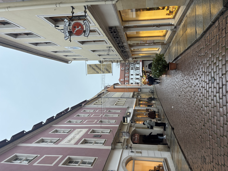 The image depicts Rabener Passage in Meissen, Germany, a narrow cobblestone street viewed looking down its length. The street surface is wet and reflective under an overcast sky.
On the left side, a multi-story building with a pale reddish-pink facade features multiple windows with white frames. The name "Rabener Passage" is vertically inscribed on its wall. At street level, an entrance displays a sign for "Rabener Keller." Two individuals are observed near this entrance: one in a dark jacket, the other in a light grey jacket. Further down the street on the left, additional buildings of varying colors, including grey and orange, recede into the distance. A shop sign for "SONJA" is visible on a building further down the left.
On the right side, a multi-story building with a pale yellow facade houses "Konditorei Café Schreiber," identified by a prominent wrought-iron hanging sign featuring a red circle with a white figure, and text indicating "Konditorei Café Schreiber." The storefront features large, brightly lit display windows. A potted green plant sits on the sidewalk in front of this building.
Overhead, decorative string lights span across the street. A large banner displaying "Sächsische Konditorei Waren" (Saxon Confectionery Goods) is suspended further down the street. In the mid-ground, a group of approximately three to five individuals walks away from the viewer along the wet cobblestone street. Additional indistinct figures are visible further in the distance. Artificial lighting emanates from the shop interiors, providing warm illumination against the subdued natural light of the overcast environment.