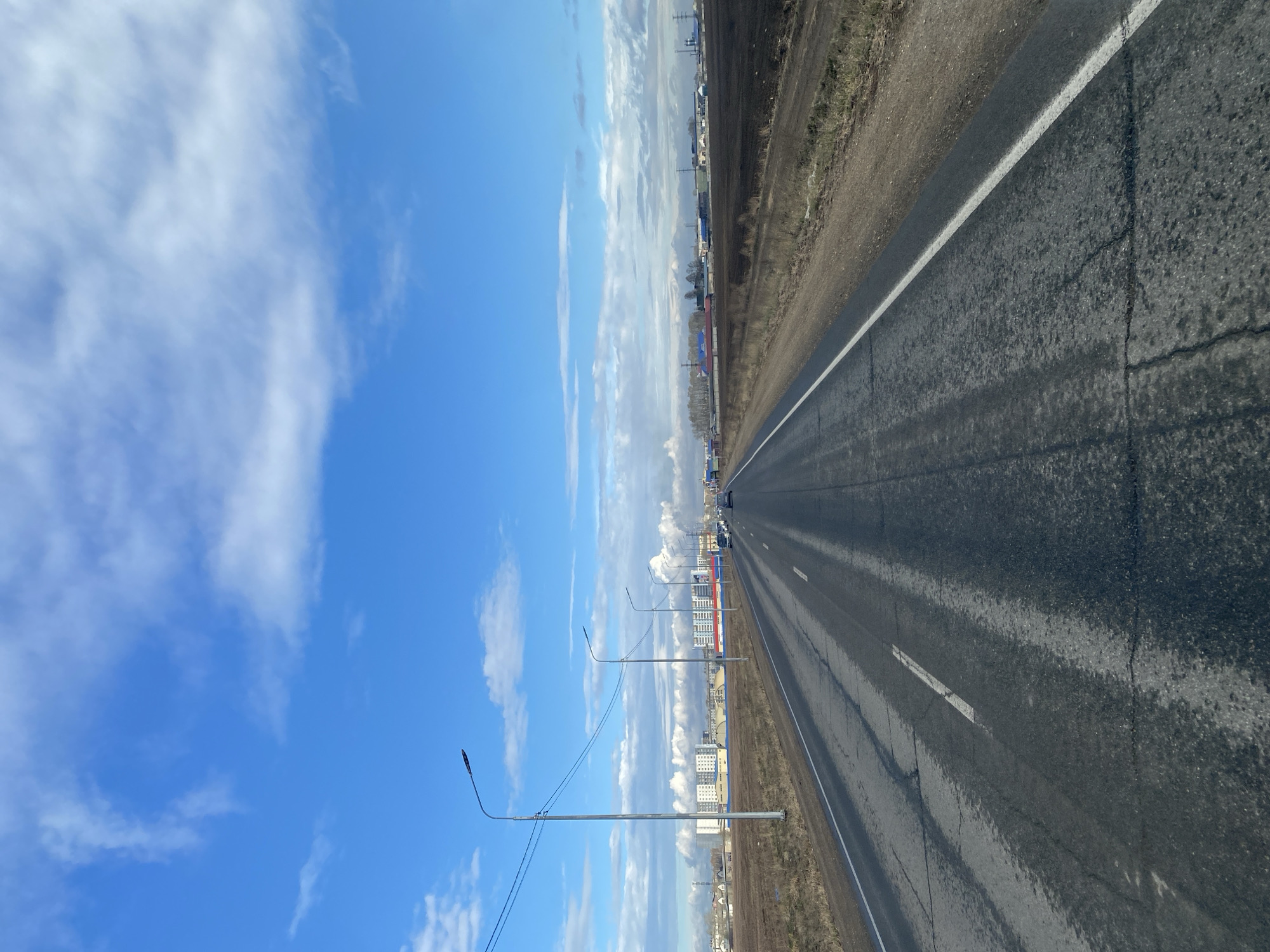 The image depicts a straight asphalt road stretching into the distance under a blue sky with scattered clouds. The road appears slightly wet, with visible reflections. To the right of the road is a dirt shoulder with vegetation. The left side features streetlights and a distant view of buildings, including what appears to be apartment complexes and industrial structures with smoke rising from chimneys. The location is identified as Usolye-Sibirskoye, Russia. There are a few vehicles in the distance along the road.