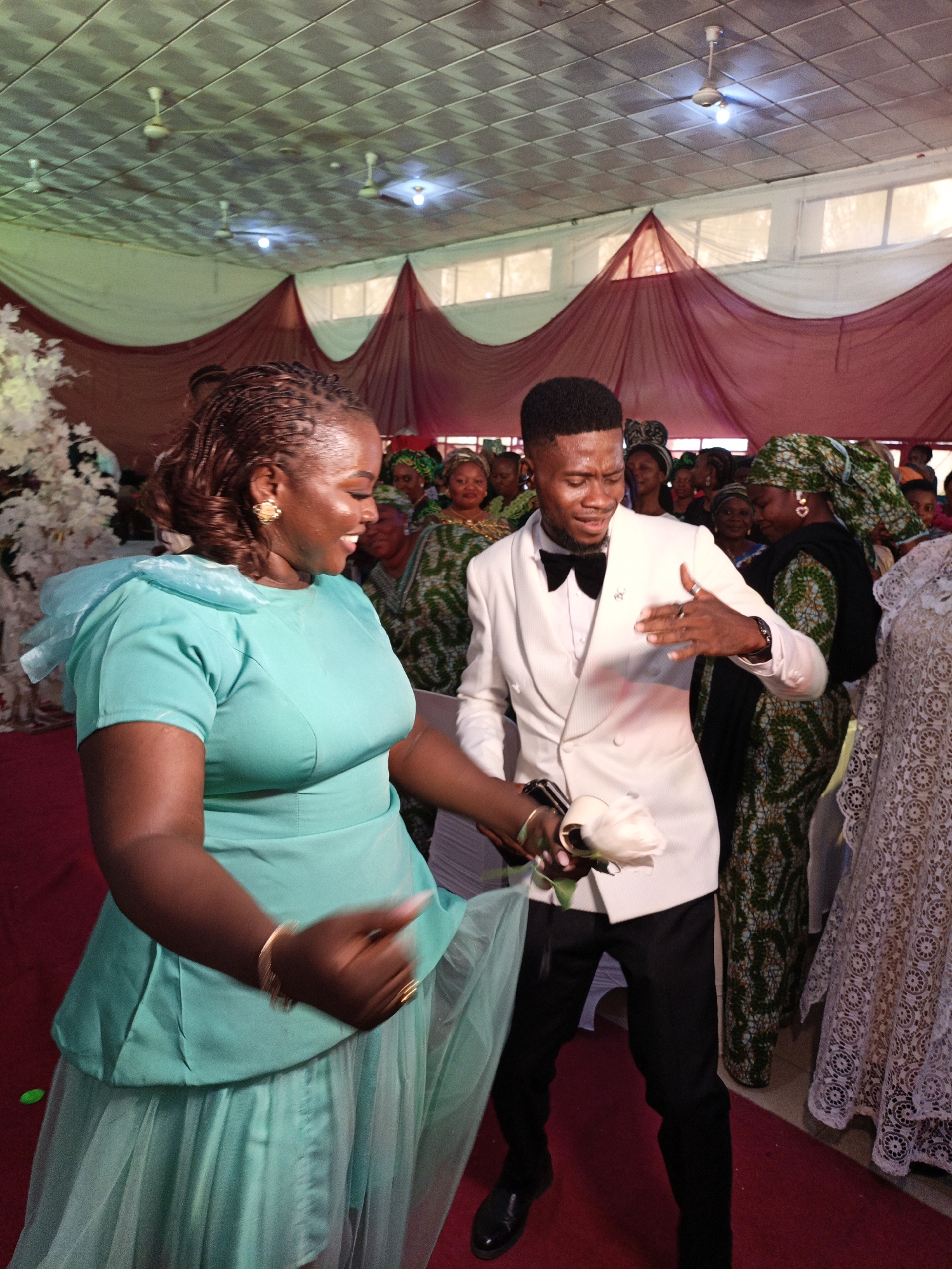 Inside a building in Fagge D2, Nigeria, a man and a woman are dancing. The woman is wearing a turquoise dress with a flowing skirt, while the man is wearing a white suit with a black bow tie. He holds a white rose. Both appear joyous. Several other people are visible in the background, dressed in various colorful attires. The building has a decorated ceiling with hanging fabric and ceiling fans. The floor is covered in a red carpet. The event appears celebratory, possibly a wedding reception.