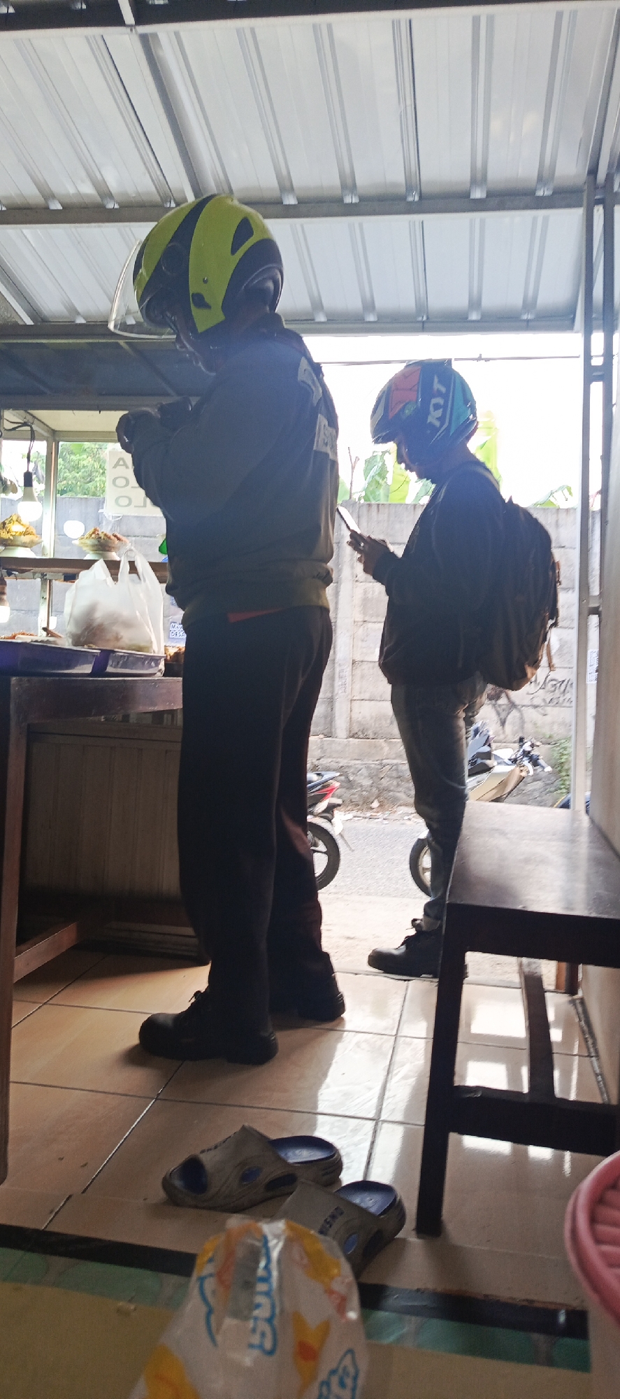 Two individuals wearing helmets are standing at the entrance of a food stall in Sukaresmi, Indonesia. The person on the left wears a yellow and black helmet and a dark long-sleeved shirt. They are holding a mobile phone. The person on the right wears a blue, orange, and black helmet and a dark jacket, also holding a mobile phone. They have a backpack slung over their shoulder. In the foreground, a plastic bag with colorful graphics is on the floor, along with a pair of gray and blue sandals. The food stall has shelves displaying food items. The flooring appears to be tiled. In the background, a motorcycle is visible. The overall setting suggests a casual outdoor environment.