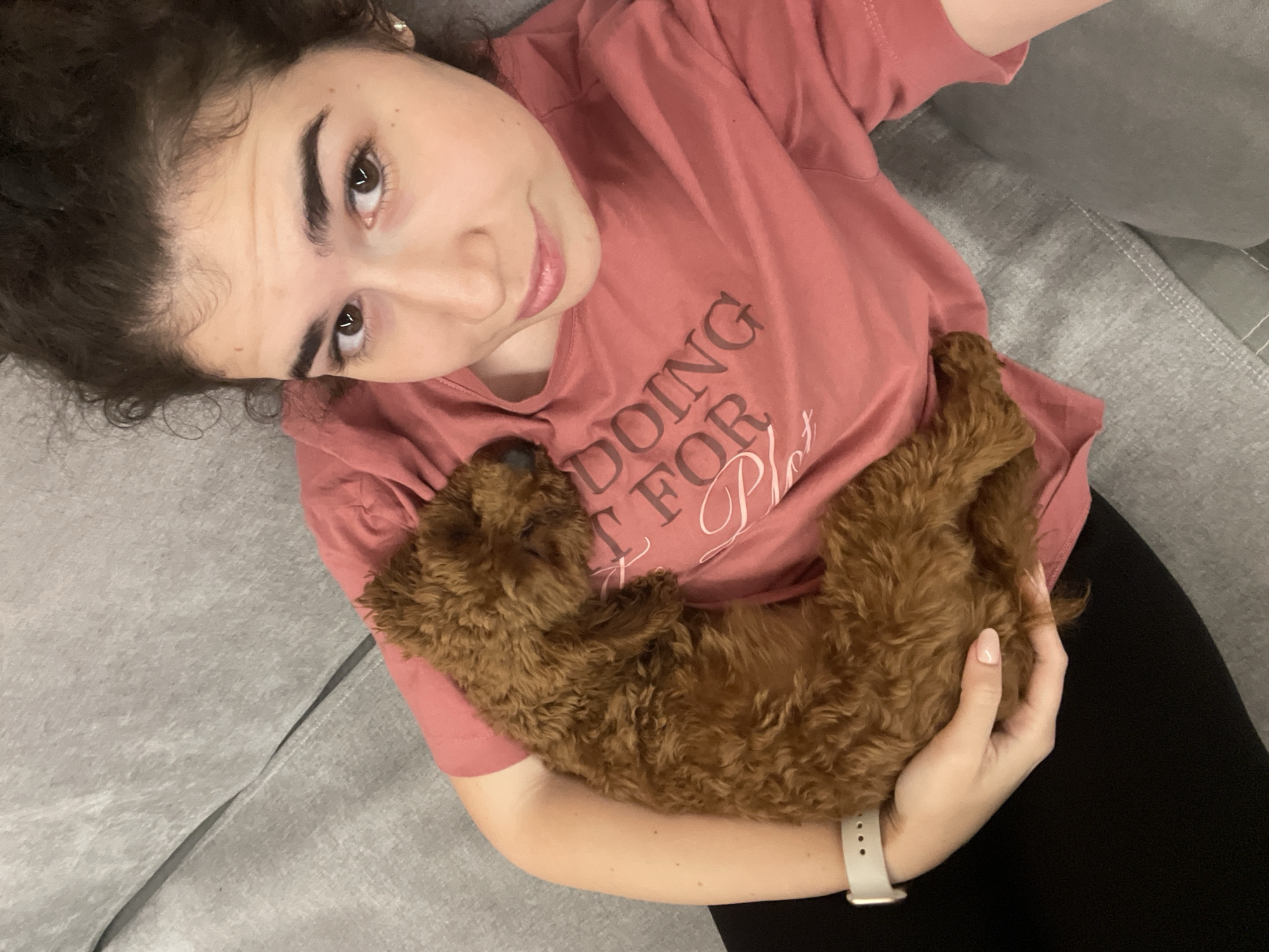 A woman with dark curly hair is lying on a grey sofa. She is wearing a pink t-shirt. A small, brown, curly-haired dog is curled up on her chest, held in her arms. The woman is looking at the camera and smiling slightly. The location is indiscernible; Sofia, Bulgaria cannot be confirmed from the visual information.