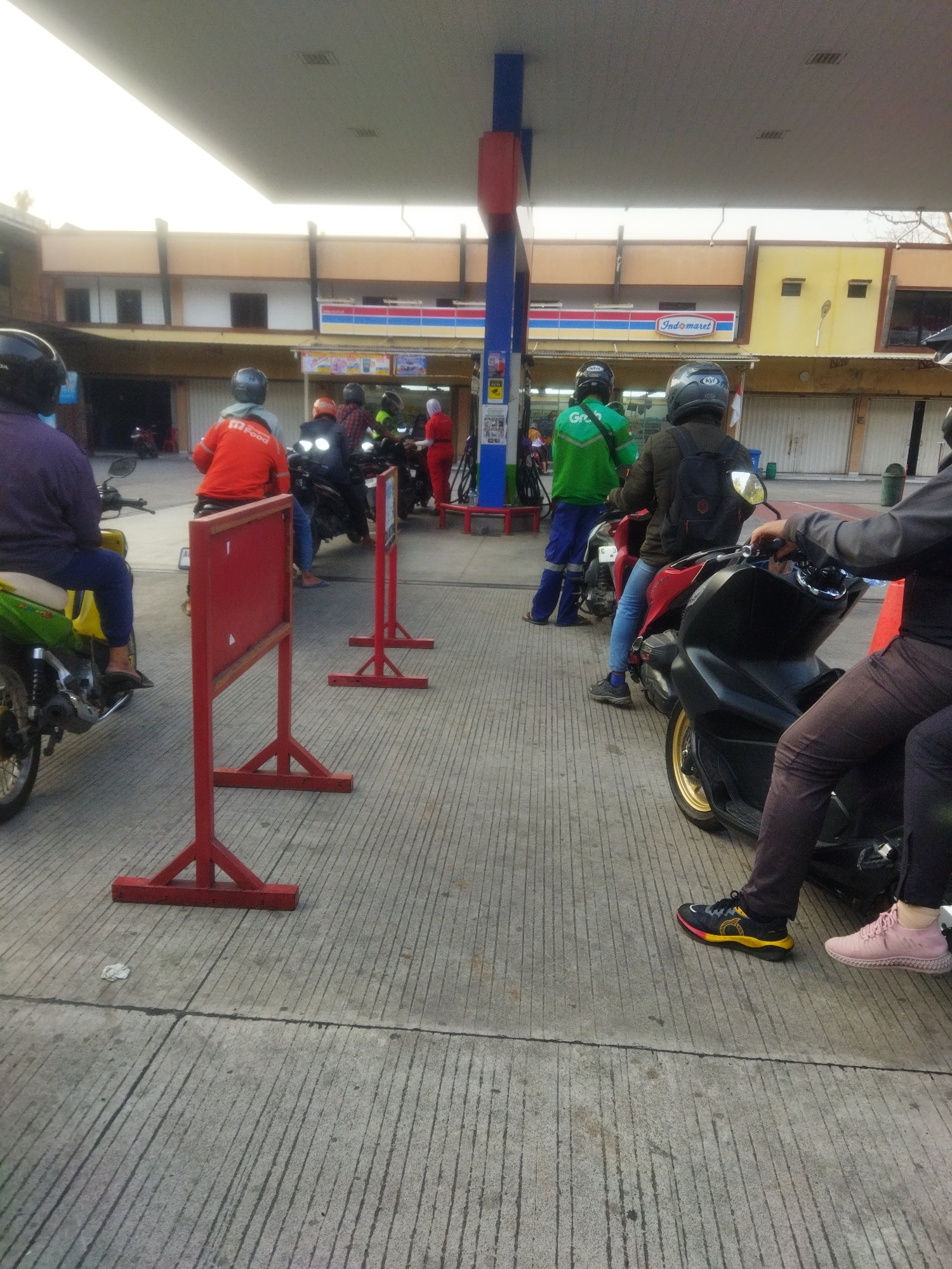 The image depicts a queue of people on motorbikes at a gas station, likely in Semarang, Indonesia, given the presence of an "Indomaret" convenience store, a common chain there. The setting is an outdoor paved area under a gas station canopy. Several individuals are seated on their motorbikes, some appearing to be waiting for service.
In the foreground, a person in dark pants and a dark jacket, wearing black and yellow sneakers, is on a black motorcycle. To their right, a person in pink sneakers is partially visible. Further into the scene, a person in blue pants and a green jacket with "Grab" branding on the back is on a motorbike. Another individual in a dark jacket and helmet is also on a motorbike.
Behind them, a person in an orange jacket with "Go Food" branding on the back is on a motorbike. There are other motorbikes and individuals in the background, suggesting a busy gas station. Red traffic barriers are placed on the pavement. The architecture of the buildings behind the gas station, including the "Indomaret" sign, provides further context about the location. The lighting suggests it is daytime, possibly late afternoon.