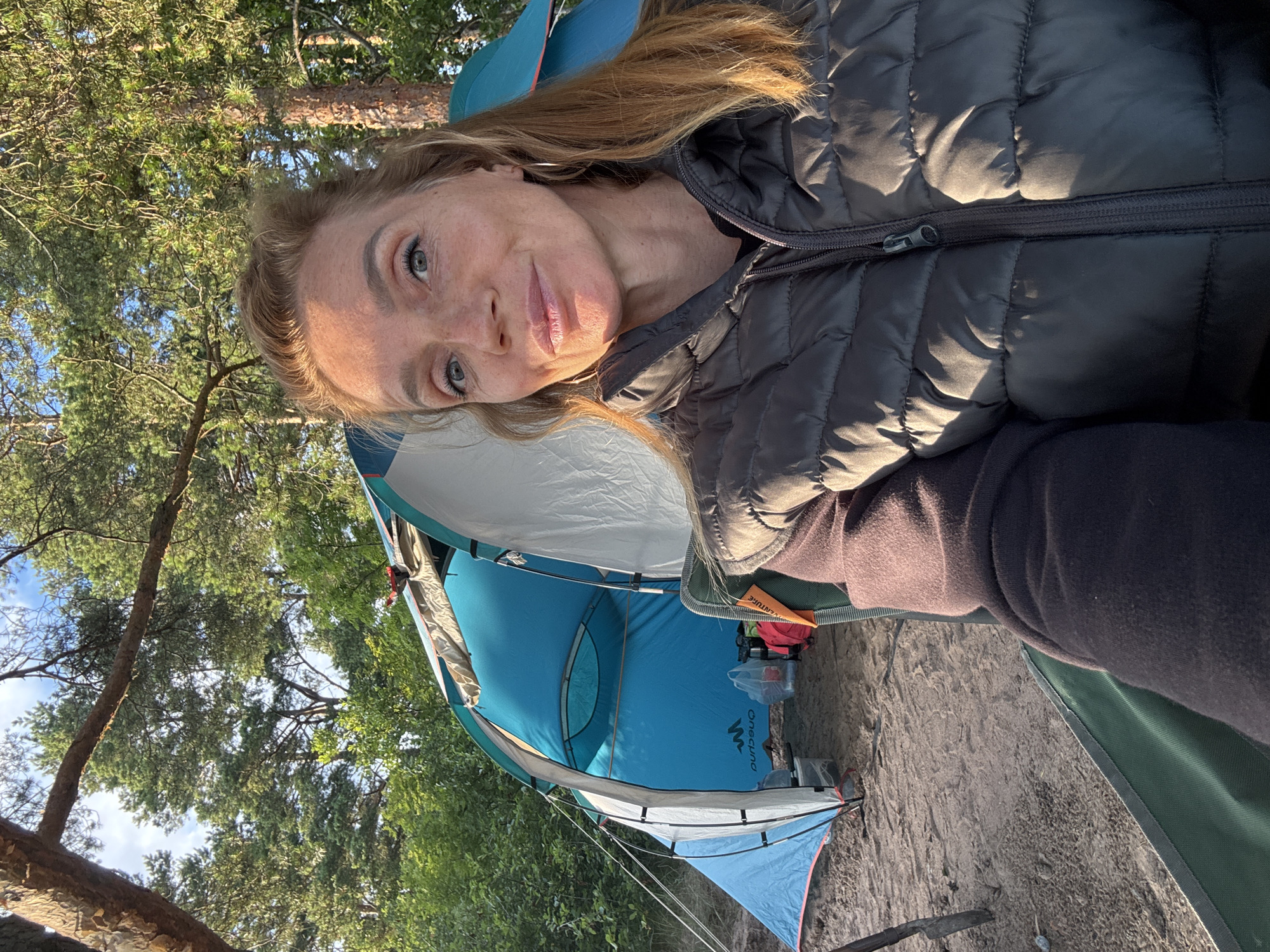 A woman is taking a selfie while sitting in a camping chair outdoors. She is wearing a dark puffer vest over a dark long-sleeved shirt and has blonde hair. Behind her, a blue tent is set up on sandy ground, surrounded by trees and vegetation, suggesting a natural environment. The lighting indicates it is daytime, possibly late afternoon or early morning, given the sun's angle. The image does not provide any specific details about the city of Shepelevo, Russia.
