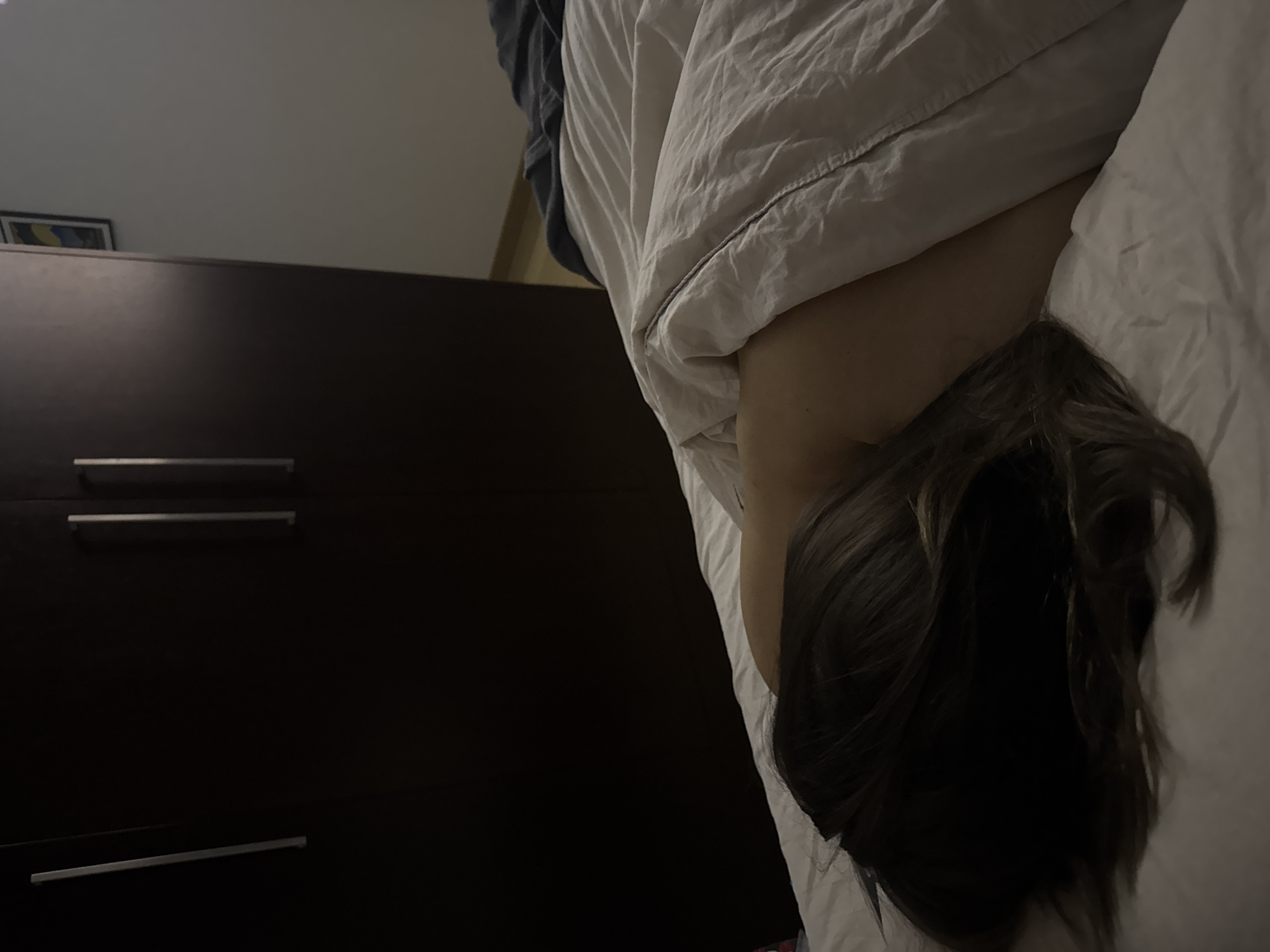 A person lies in bed covered by white blankets, with only their head and upper back visible. The head has dark hair and is turned away from the camera. To the left of the bed is a dark brown dresser with silver handles. The setting is a bedroom, likely indoors. Location is unknown.