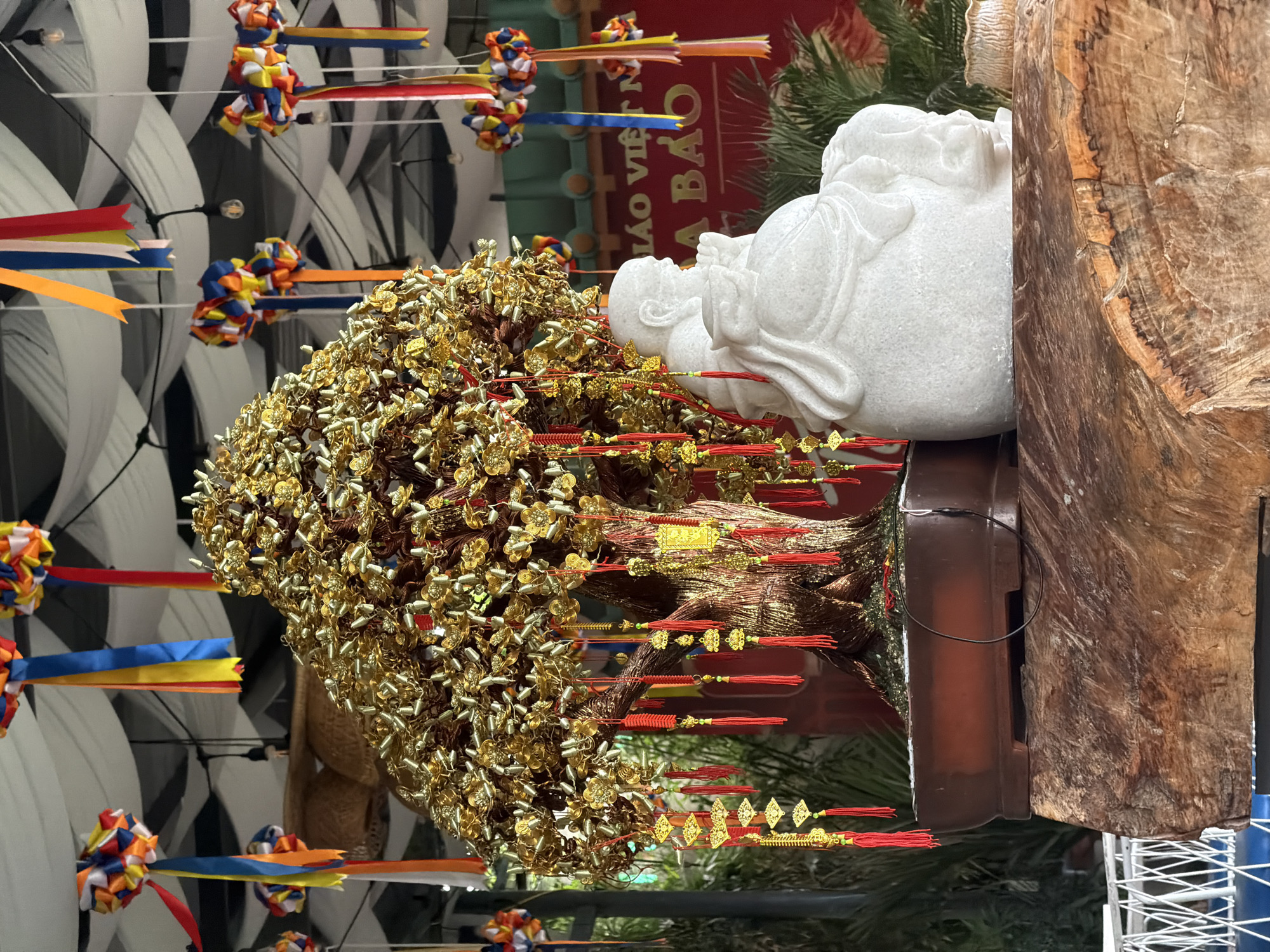 The image shows a statue of a reclining figure in white marble, juxtaposed with a tree-like sculpture adorned with golden embellishments and red tassels. The marble sculpture rests atop a dark brown base, which is adjacent to a weathered wooden pillar. Multi-colored ribbons and decorative ornaments are suspended from the ceiling in the background, and some Vietnamese signage is visible behind the plants. The location is Bac Nha Trang Ward, Vietnam.