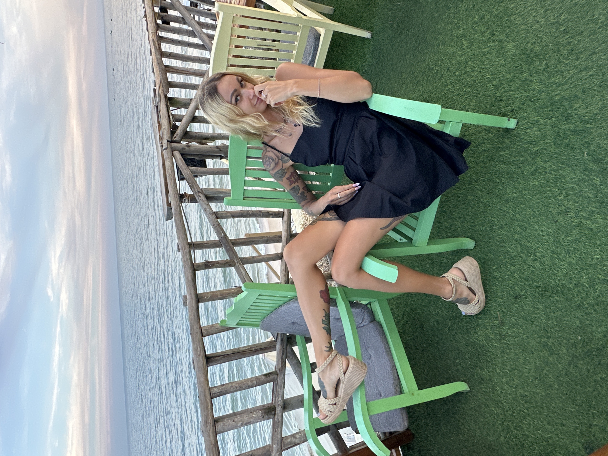 A young Caucasian woman with blonde hair is seated on a green chair outdoors, facing the camera. She is wearing a black slip dress and tan wedge sandals. Her left leg is extended, and her right hand is touching her face. She has tattoos on her arms and legs. The chair is positioned on a patch of green astroturf, next to another green chair and a light-colored bench. Behind her is a wooden railing overlooking the sea and a cloudy sky. The setting appears to be a waterfront establishment in Phu Thuy Ward, Vietnam.