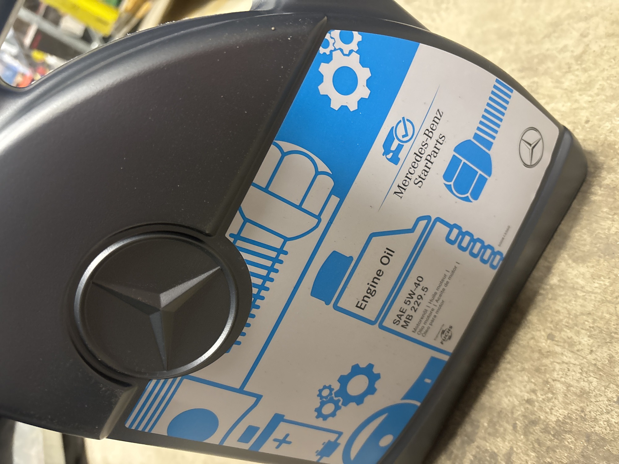 A close-up, angled shot shows a dark gray plastic bottle of Mercedes-Benz StarParts engine oil. The label is predominantly white with blue graphics and text. Prominently displayed is the Mercedes-Benz three-pointed star logo embossed on the bottle. The label features several blue icons, including a car with a checkmark, gears, and abstract shapes. Text on the label indicates "Engine Oil," "SAE 5W-40," and "MB 229.5." The words "Motoröil | Huile moteur | Olio motore | Aceite de motor | Óleo para motor" are also visible. Below this, a smaller logo reads "FUCHS." The bottle is resting on a light-colored, textured surface that appears to be a floor or countertop. The background is blurred, but hints of shelves and other objects suggest an indoor environment. The overall impression is one of a product detail shot.