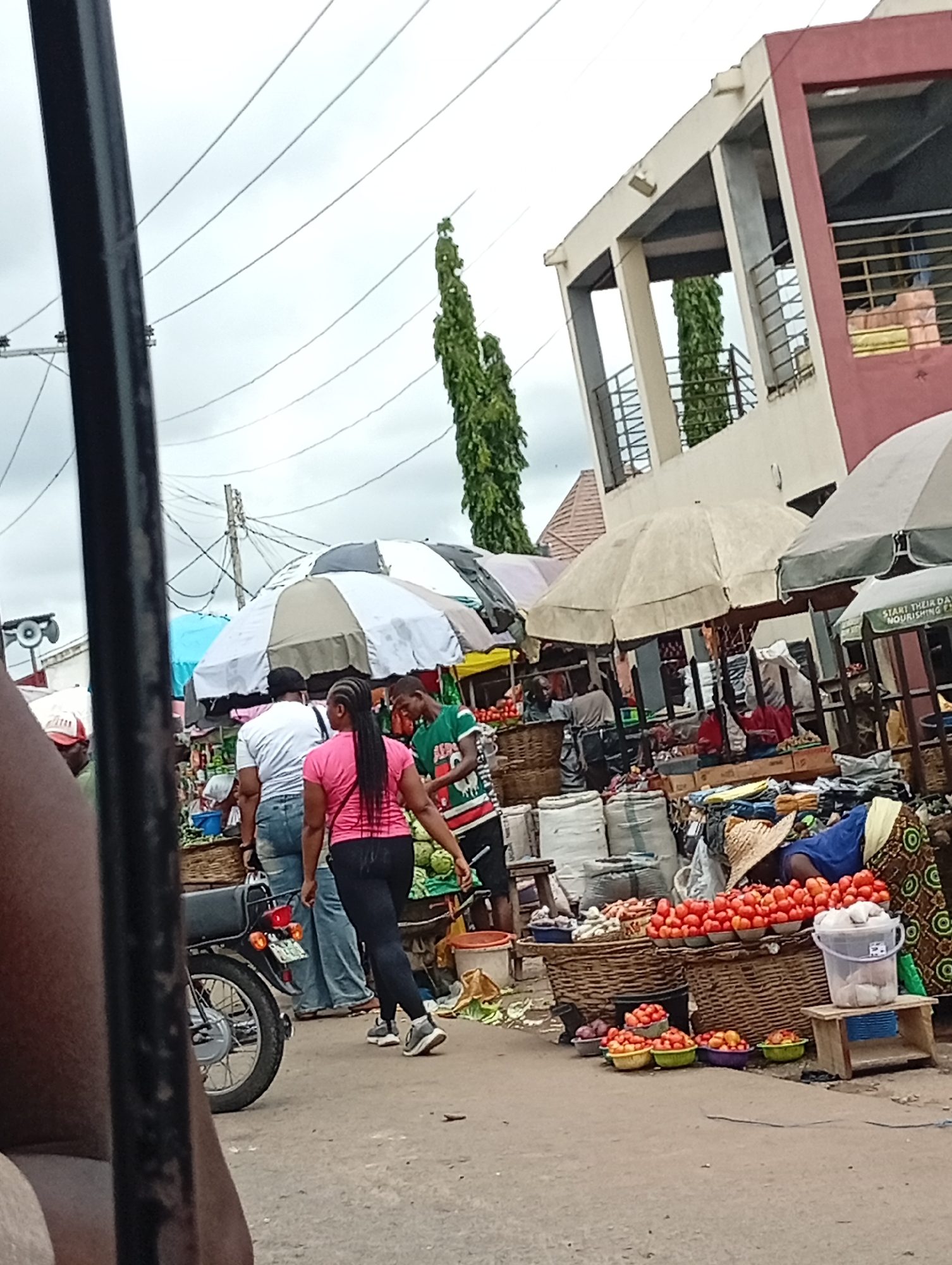 The image depicts a bustling market scene in Lokoja, Nigeria. A woman with long, dark hair wearing a pink shirt and black leggings walks through the market. To her left are a man in a white shirt and jeans, and a parked motorbike. In front of her, a man in a green shirt inspects produce. Stalls covered by umbrellas line the street, displaying tomatoes, onions, and other goods in baskets and bags. A woman wearing a straw hat leans over a display of tomatoes. In the background, a two-story building with open walkways and a red and beige color scheme is visible. Power lines crisscross the sky, which is overcast. The foreground includes a blurry vertical black bar on the left side of the frame.