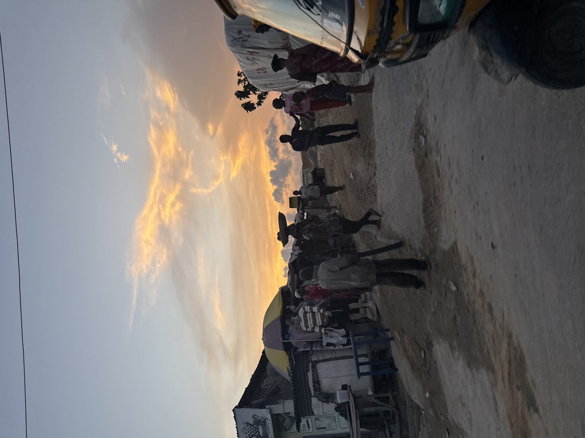 The image captures a street scene in Ado, Nigeria, during sunset or twilight. In the foreground, there's a glimpse of a yellow auto rickshaw (tuk-tuk). The middle ground shows a group of people walking and interacting on a dirt road or pathway. Structures, possibly shops or stalls, line the road on one side, including one with a yellow and purple umbrella. The background is dominated by a sky with a vibrant orange and yellow sunset. The overall image appears to be captured at ground level, providing a pedestrian's perspective of the everyday life in Ado.