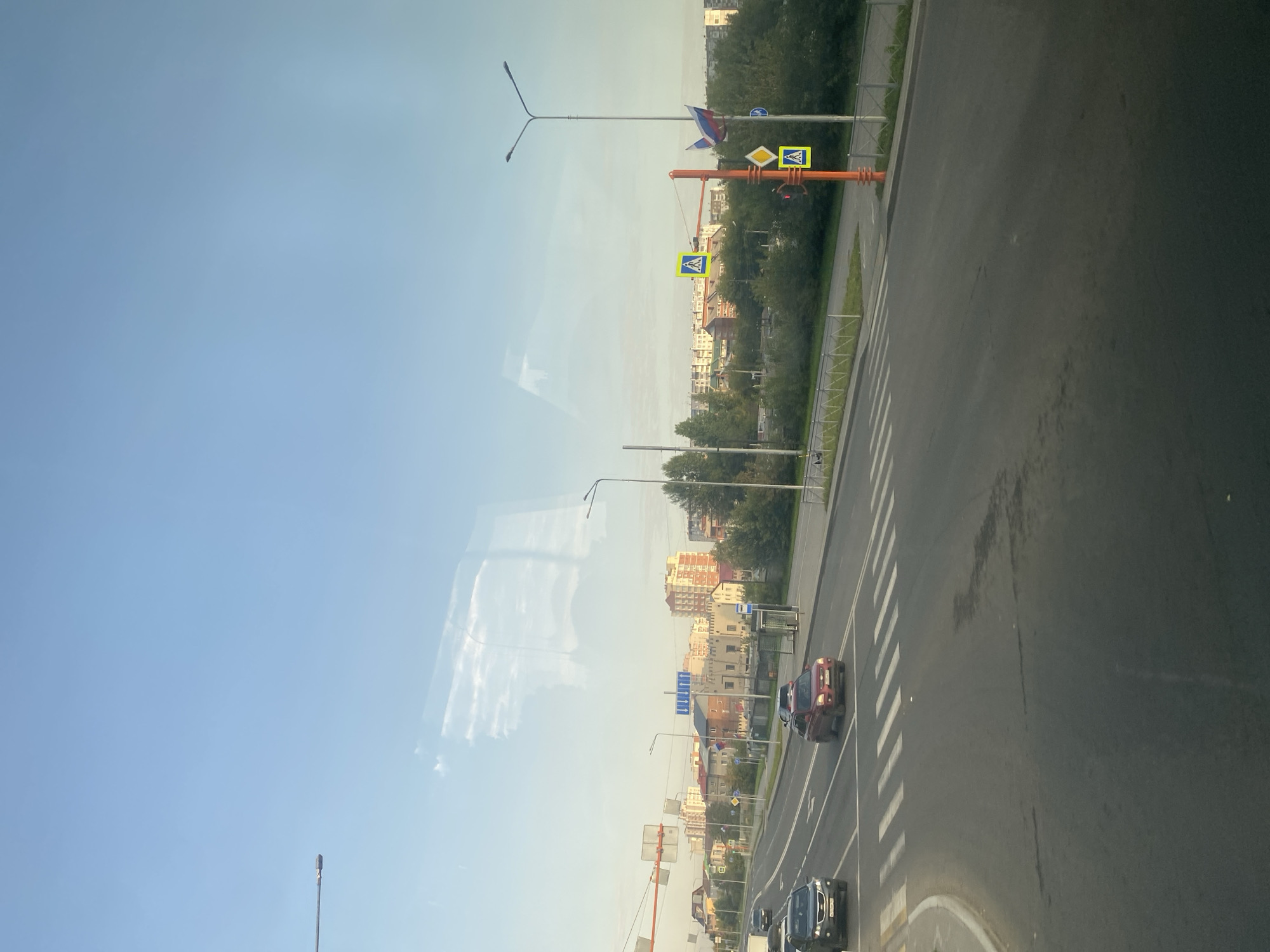 The image depicts a wide, multi-lane asphalt road in Kemerovo, Russia, during daylight. The sky is a clear, pale blue, with some white cloud reflections visible. The road features a pedestrian crossing marked by white stripes. Several cars are on the road: a dark SUV in the left lane, a red SUV in the right lane approaching the pedestrian crossing, and a black car further ahead. To the right of the road, there is a sidewalk with a bus stop shelter and a traffic light pole adorned with pedestrian crossing signs. Further in the background, a row of apartment buildings of varying heights and architectural styles, some with visible balconies and windows, line the horizon. Trees and green foliage are visible between the road and the buildings, indicating a mix of urban and natural elements. Russian flags are displayed on a pole near the pedestrian crossing signs. The overall scene suggests a typical urban environment in Russia during a calm, clear day.