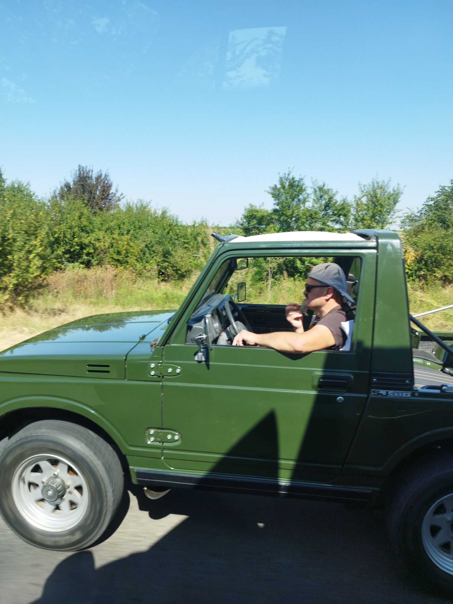 A green, open-top Suzuki Samurai SJ410 is driving on a paved road. The driver, a man wearing a grey baseball cap and sunglasses, is visible from the side, with his left arm resting on the open window. He appears to be holding something small to his mouth. The vehicle is surrounded by green foliage and trees, indicating a rural or natural setting. The sky above is clear and blue. The image was taken in the vicinity of Cocani, Romania.