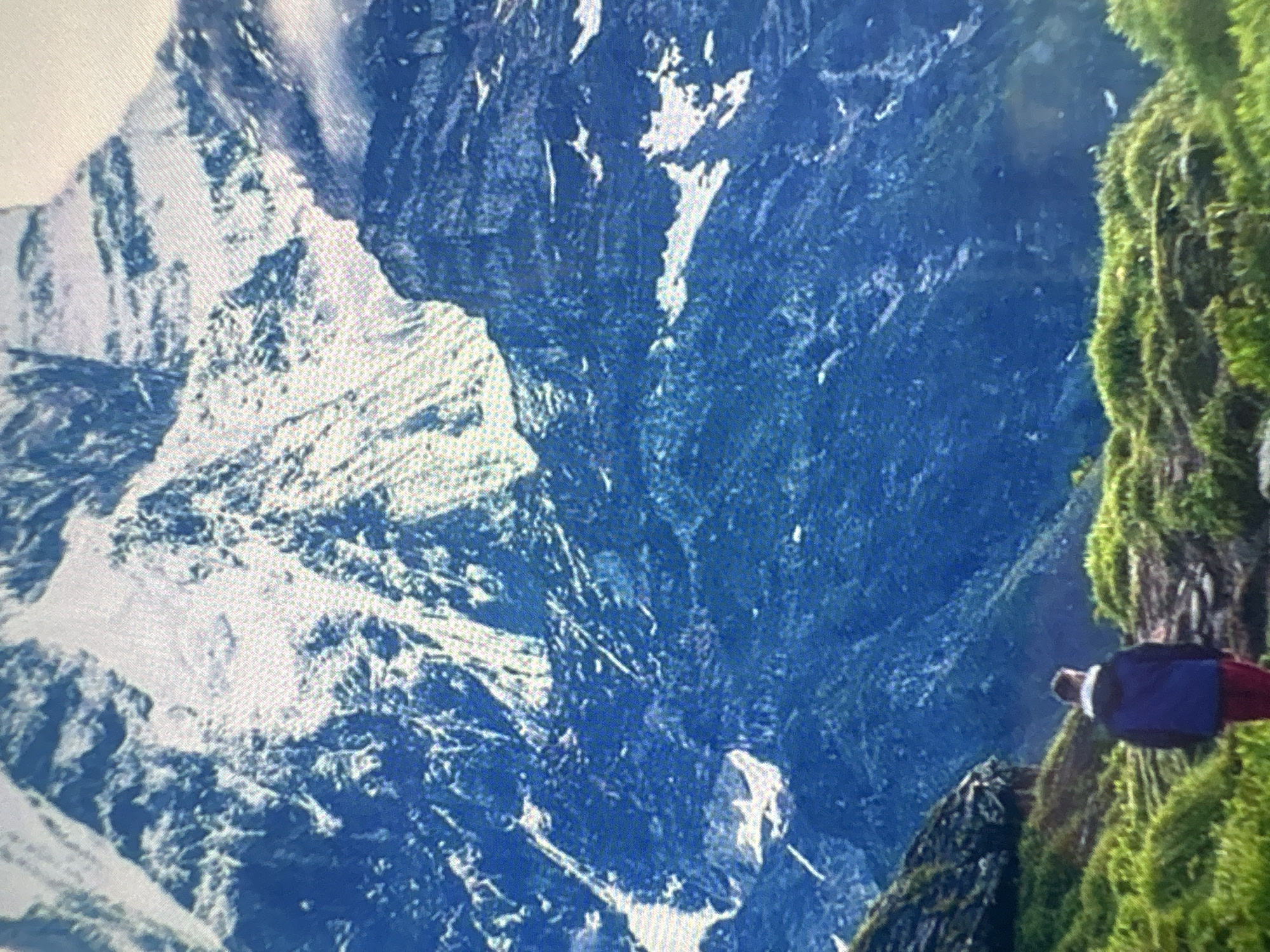 A landscape view of mountainous terrain, primarily in shades of blue and white, resembling snow-covered peaks and rocky slopes. On the right side of the image, green vegetation, possibly moss or grass, clings to a cliff. A figure, appearing to be a person, is situated near the edge of the cliff, partially obscured by the vegetation. They are wearing clothes in dark blue, white and red. The perspective is from above, looking down towards the valleys below. This image doesn't have enough information to pinpoint the location specifically in Kukorevo, Bulgaria or have any interactions.
