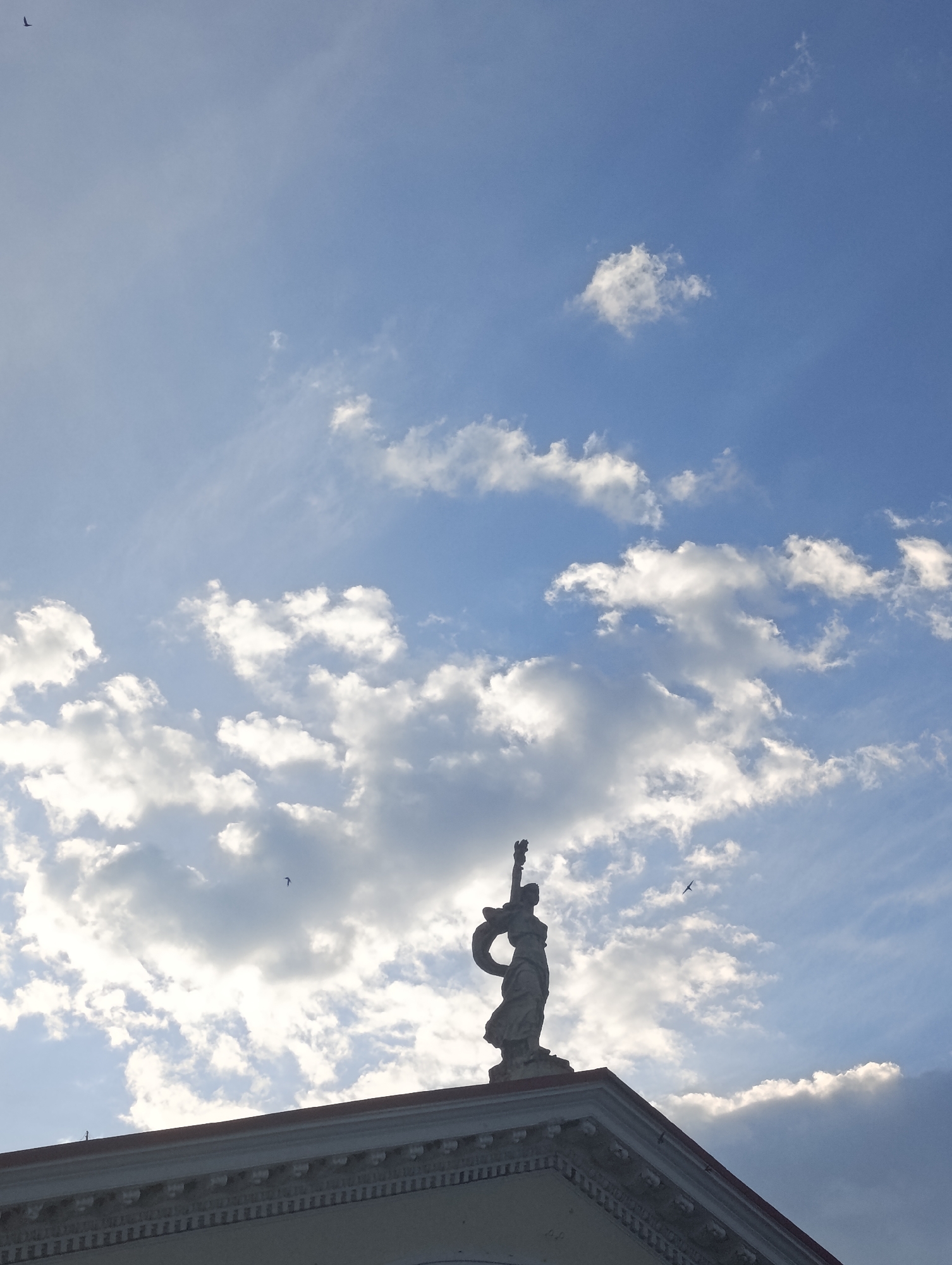 A silhouetted statue of a woman holding a torch stands atop a building against a blue sky with scattered clouds. The statue is positioned on the right side of the frame, and the building's roof extends across the bottom. Several birds are visible in the sky, appearing as small dark shapes. The lighting suggests it is either sunrise or sunset, with the sun partially obscured by clouds, creating a bright glow.