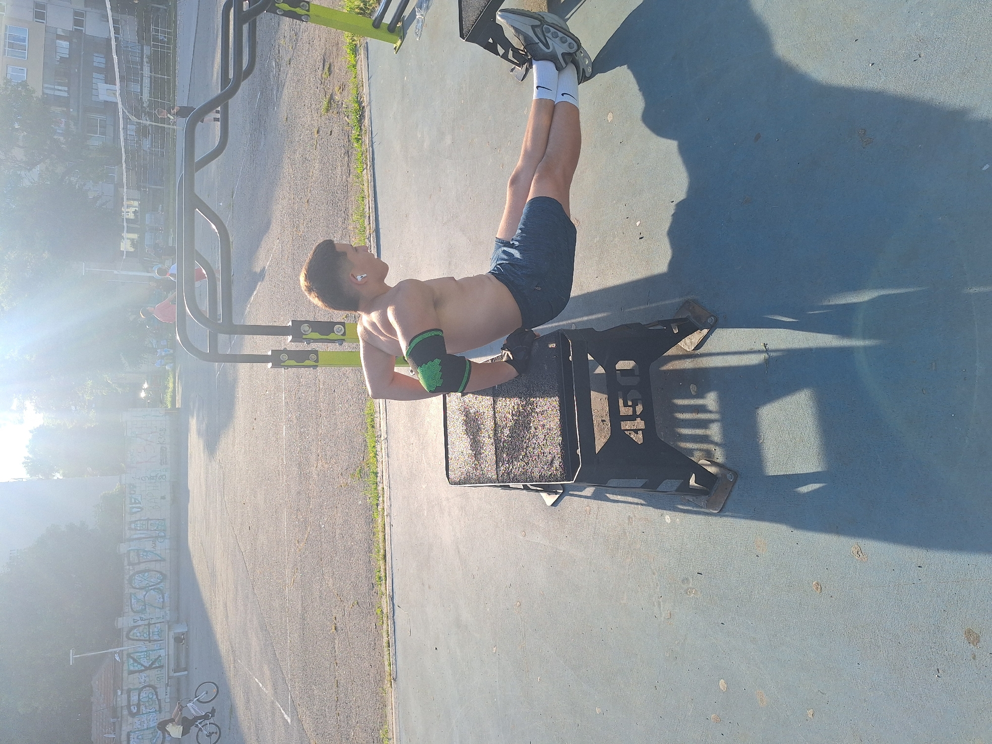 In Varna, Bulgaria, a young man is performing triceps dips on a black plyometric box at an outdoor gym. He is shirtless and wearing blue athletic shorts, green elbow sleeves, white socks, and grey athletic shoes. His legs are extended forward, and his hands are gripping the edge of the box. The gym equipment is located on a blue-colored paved surface. In the background, a volleyball net is visible, along with some trees and buildings. There are other individuals in the background, including a person riding a bicycle and some children playing. The scene is bathed in bright sunlight, casting long shadows on the ground. The overall environment appears to be a public recreational area.