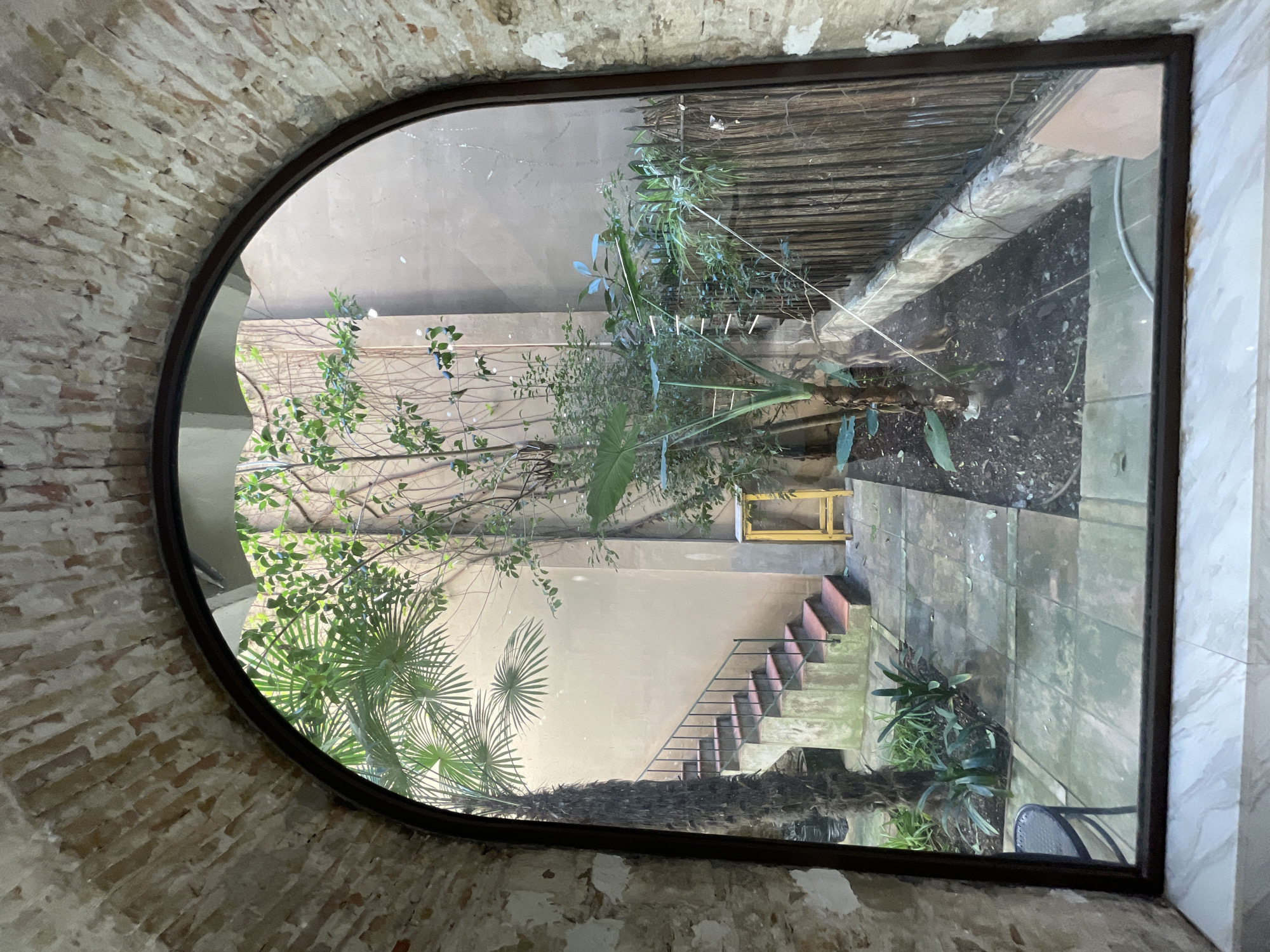 The image captures an interior view looking out through a large, arched window into a vibrant outdoor courtyard or garden. The interior wall surrounding the window arch is made of light-colored, weathered bricks, suggesting an old or rustic setting. A dark, possibly metal, frame outlines the window's arch.
The outdoor scene is dominated by varied plant life and architectural features. On the left and in the foreground, lush green foliage includes distinctive fan-shaped palm leaves and other leafy trees. A light-colored wall forms the main background of the courtyard. A set of concrete stairs with reddish-brown treads and a dark metal handrail ascends along the right side of this wall. Near the top of the stairs, a small, bright yellow box-like structure is visible.
The ground level of the courtyard consists of both paved sections with square tiles and areas of dark soil where smaller plants and ground cover grow. Towards the upper right, a wooden slatted fence or screen is visible. A dark metal outdoor chair is partially seen in the lower right corner of the courtyard.
The scene is well-lit by natural daylight, indicating it is daytime with clear weather conditions. No people are present, and no specific activity is taking place. The courtyard appears to be a peaceful, green space, reflecting a Mediterranean climate consistent with Barcelona. No text is visible in the image.