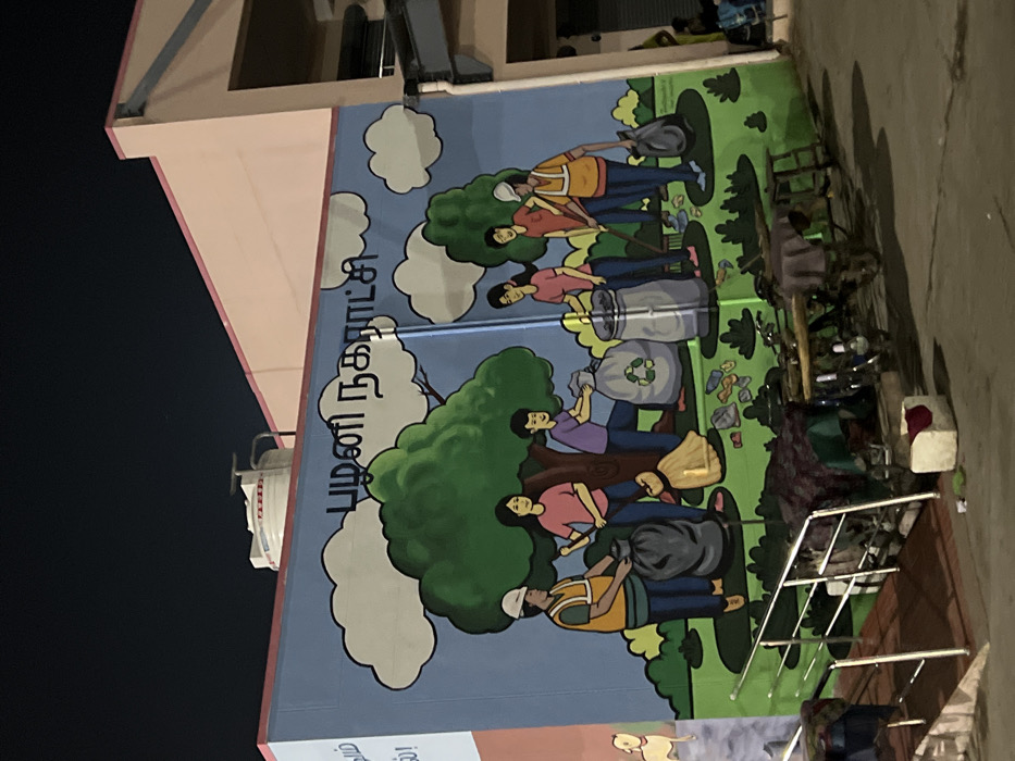 The image features a large, colorful mural painted on the side of a building, depicting a scene focused on cleanliness and waste management. The mural, oriented vertically along the building's facade, illustrates several cartoon-style individuals engaged in various cleaning activities. These figures, including both men and women, are shown sweeping the ground with brooms, picking up litter, and disposing of waste into garbage bins and bags, some of which feature recycling symbols. The background of the mural consists of green grassy areas, trees, and white clouds against a light blue sky, portraying an ideal clean environment. Scattered trash items, such as bottles and wrappers, are also depicted on the ground within the mural, representing the problem being addressed.
Visible in Tamil script on the mural is "பழனி நகராட்சி" (Palani Nagaratci), which translates to "Palani Municipality," indicating the local government body responsible for the message. Further down, partially visible text likely reads "அனைவரும் வாருங்கள்!" (Anaivarum Vaarungal!), meaning "Everyone, come!", suggesting a community call to action.
The real-world setting shows a dark, likely night sky to the left and top of the building, contrasting with the bright imagery of the mural. The building itself has light pinkish-beige walls and parts of its roofline are visible. On the ground level to the right of the mural, there is some clutter, including a bicycle, possibly some discarded items or furniture, and a metal railing. The ground surface appears to be paved. The mural serves as a public awareness message in Palani, India, promoting civic responsibility and a clean environment.