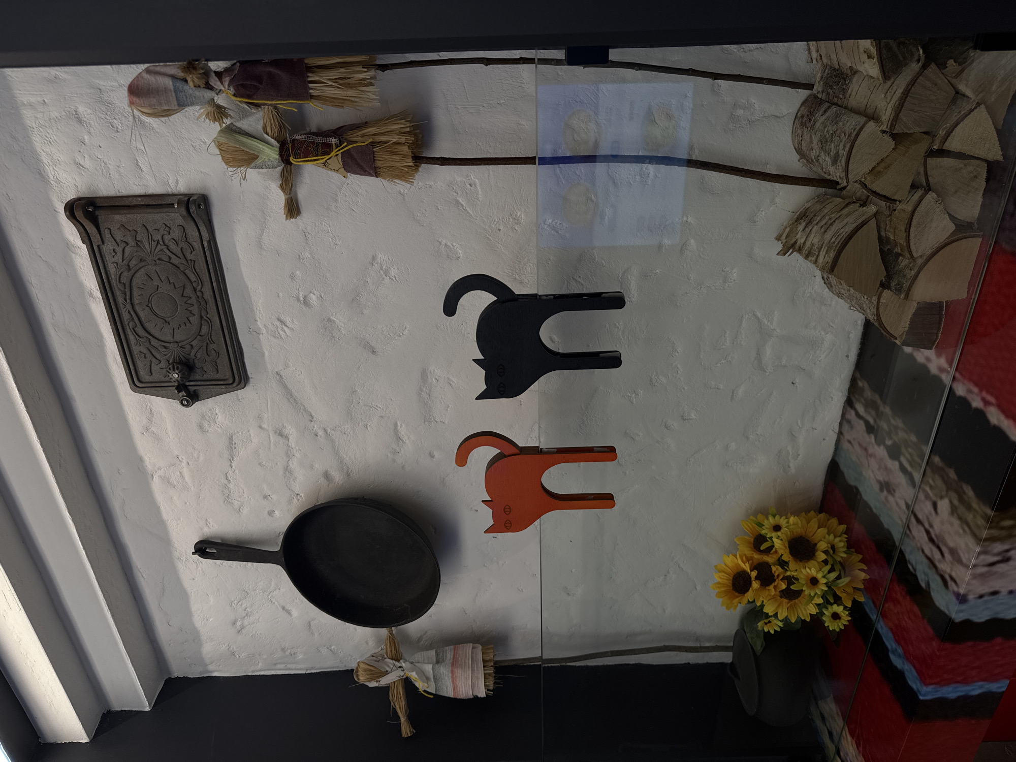 This image presents an upward-facing view of a white, textured wall adorned with various decorative and utilitarian items, creating a rustic or traditional aesthetic. A large, vertical glass panel centrally divides the composition, reflecting parts of the objects and showing a distinct horizontal blue line.
On the left side of the wall, a dark, ornate cast iron plate or small door, featuring an intricate, symmetrical design resembling a sun or flower, is mounted. Below it, a black cast iron frying pan hangs. To the far left, several white, ridged architectural elements, possibly beams or parts of a ceiling structure, are visible.
Several small straw dolls or decorative brooms, tied with colorful fabrics, are arranged around the scene: one in the top left corner, one below the cast iron pan, and two positioned horizontally above the glass panel.
Two stylized cat figures, appearing to be cut from wood or similar material, are mounted on the wall to the right of the cast iron objects. One is black and positioned higher, while the other is orange and sits below it.
In the bottom right corner, a vase filled with bright yellow sunflowers, featuring dark centers, adds a vibrant splash of color. Above the sunflowers, a stack of cut firewood, predominantly birch, is neatly arranged. The overall texture of the wall is rough and uneven, suggesting plaster or whitewashed brick. Dark, solid surfaces define the very top and bottom edges of the frame. The scene likely depicts an interior space in Saint Petersburg, Russia, showcasing elements of local decor or folk art. No discernible text is visible.