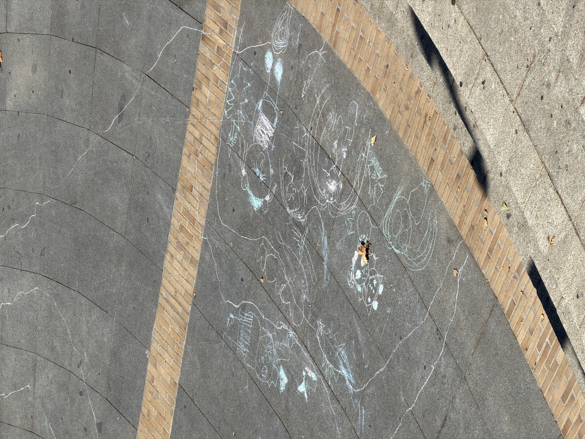The image shows a section of a paved walkway in Las Rozas de Madrid, Spain, with various chalk drawings scattered across it. The walkway is made of dark grey rectangular tiles with lighter grey grout lines. A decorative border of tan bricks runs horizontally across the middle of the frame. The chalk drawings are primarily in white and light blue, depicting a variety of childlike figures and abstract shapes. Some of the drawings appear to be faces, while others are more stylized representations of animals or objects. A single brown leaf rests on the pavement among the drawings. The scene suggests a public outdoor space, possibly a park or plaza, where children have been drawing with chalk. The light suggests it is daytime.
