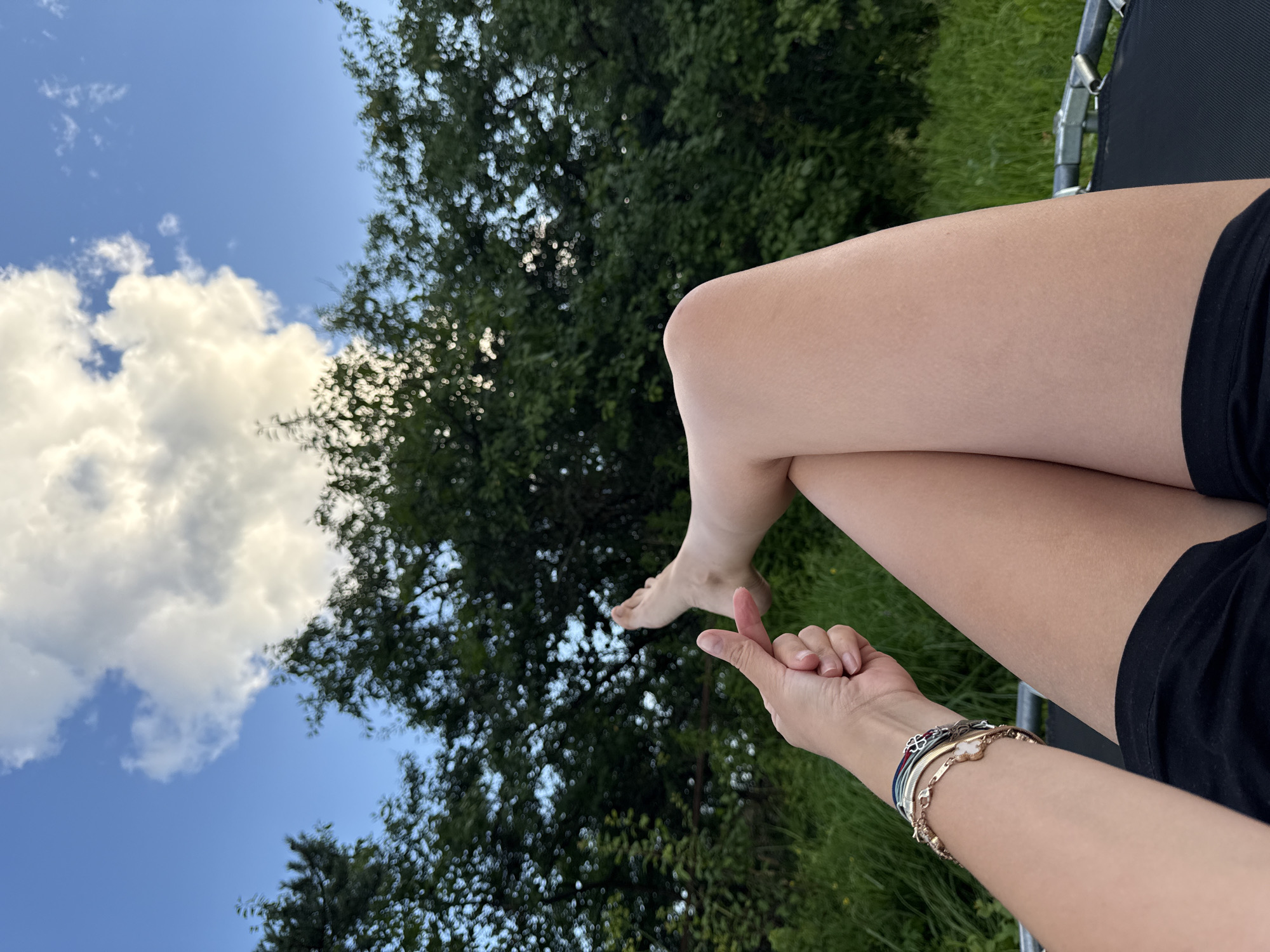 The image shows a person's legs and arm, with their legs crossed and one foot extended upwards. The person is wearing black shorts and several bracelets on their wrist. They are lying on a trampoline, with a background of green trees and a blue sky with white clouds. The setting appears to be outdoors in a rural or suburban area. The specific location in the city of Vetluzhsky, Russia, is not discernible from the image.