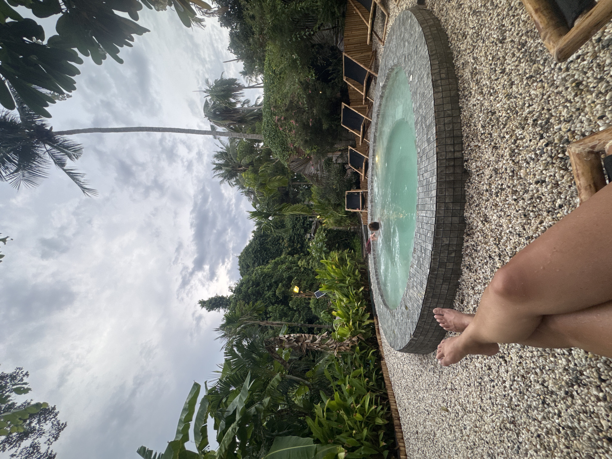 The image shows a round hot tub located in Baan Sri Thanu, Thailand. The tub has gray tiles and is surrounded by small pebbles. There are lounge chairs with dark cushions placed next to the tub, amongst lush tropical vegetation including palm trees and banana plants. One person is soaking in the hot tub and another is sitting with their legs crossed at the edge of the tub. The sky is overcast with gray clouds.