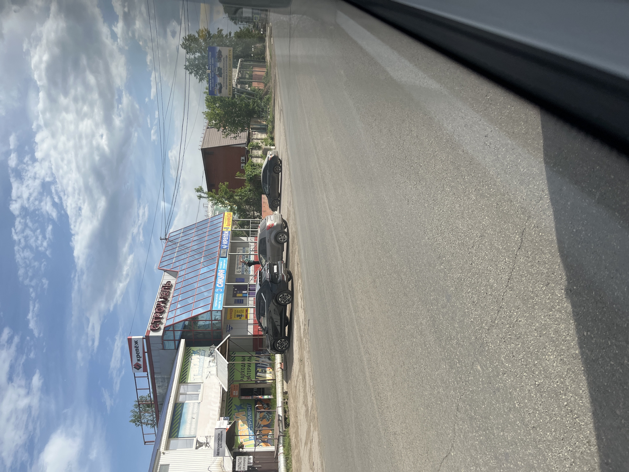 The image shows a street scene in what appears to be a Russian city. The angle suggests the photo was taken from inside a vehicle. The focus of the image is a building identified as "STROMEN," a construction and hardware store. The storefront displays signage in Russian, including "KREPEZH" and "AVTOZAPCHASTI DLYA INOMAROK", which translates to "fasteners" and "auto parts for foreign cars", respectively. Advertising for "SAKURA" with a phone number is also visible. Three cars are parked along the street in front of the building. The sky is partly cloudy. The address is located in the city of Chaikovsky, Russia.