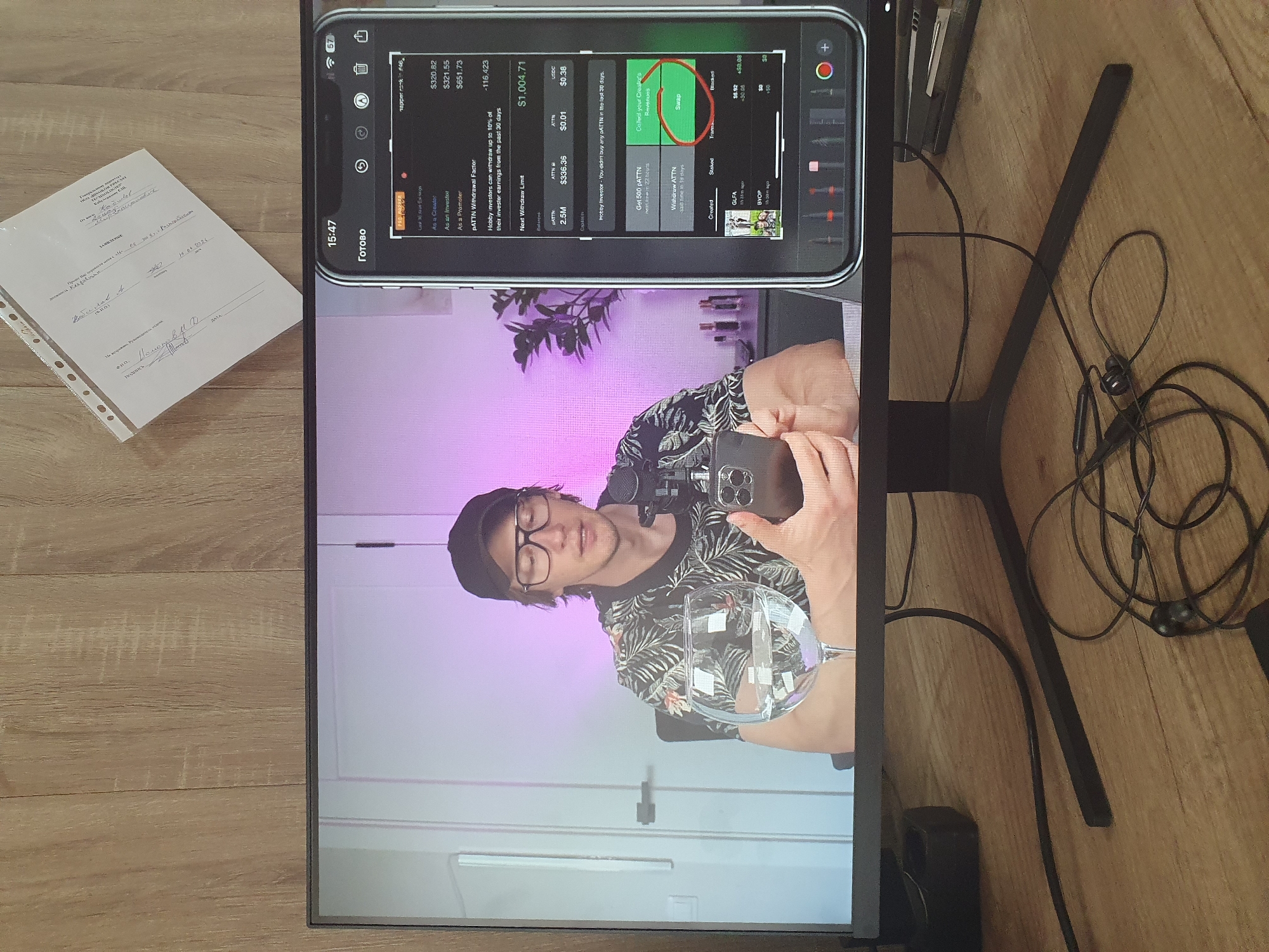A medium shot captures a person wearing glasses and a floral shirt, sitting in front of a computer monitor that displays a video feed of themselves. They hold a smartphone in their right hand, looking down at it. The monitor screen also shows a separate app interface, possibly related to cryptocurrency or digital assets. To the right of the monitor, a document with handwritten text and stamps is partially visible on a wooden desk. The background consists of a light-colored wall with a door and a plant. The overall setting appears to be an indoor workspace or studio. There is no information in the image to determine the location in Tashkent, Uzbekistan.