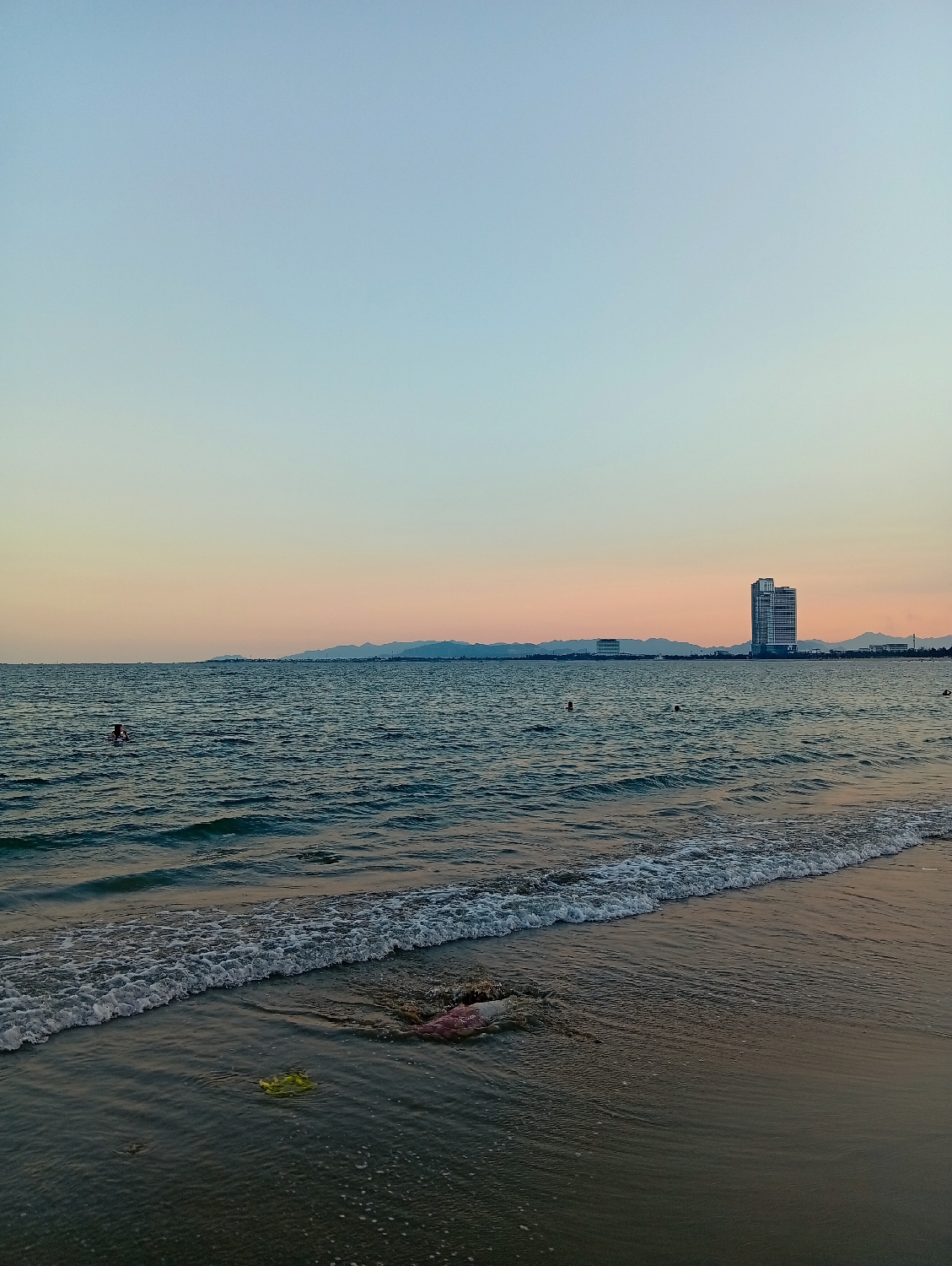 This media file captures a serene coastal scene in Khánh Hải, Vietnam, likely during the late afternoon or early evening. The sky displays a gentle gradient, transitioning from a clear light blue at the top to soft orange and pink hues along the horizon, suggesting either sunrise or sunset.
The dominant feature is the vast expanse of the ocean, which appears a deep blue-green with small, frothy waves breaking onto a sandy shoreline. In the distance, a low mountain range or hills stretches across the horizon, behind which several modern high-rise buildings stand tall, indicating a developed coastal area.
In the mid-ground, at least two individuals are visible in the water, appearing to be swimming or wading. Closer to the foreground, where the waves meet the wet sand, some litter is unfortunately noticeable. A distinct bright yellow object, possibly a plastic bag or piece of fabric, and a pinkish-purple textile-like item are caught in the shallow water near the wave line.
The atmosphere is generally calm, with no harsh weather conditions apparent. The smooth water and clear sky contribute to a peaceful ambiance, contrasting slightly with the visible man-made debris on the shore. There is no visible text in the image.