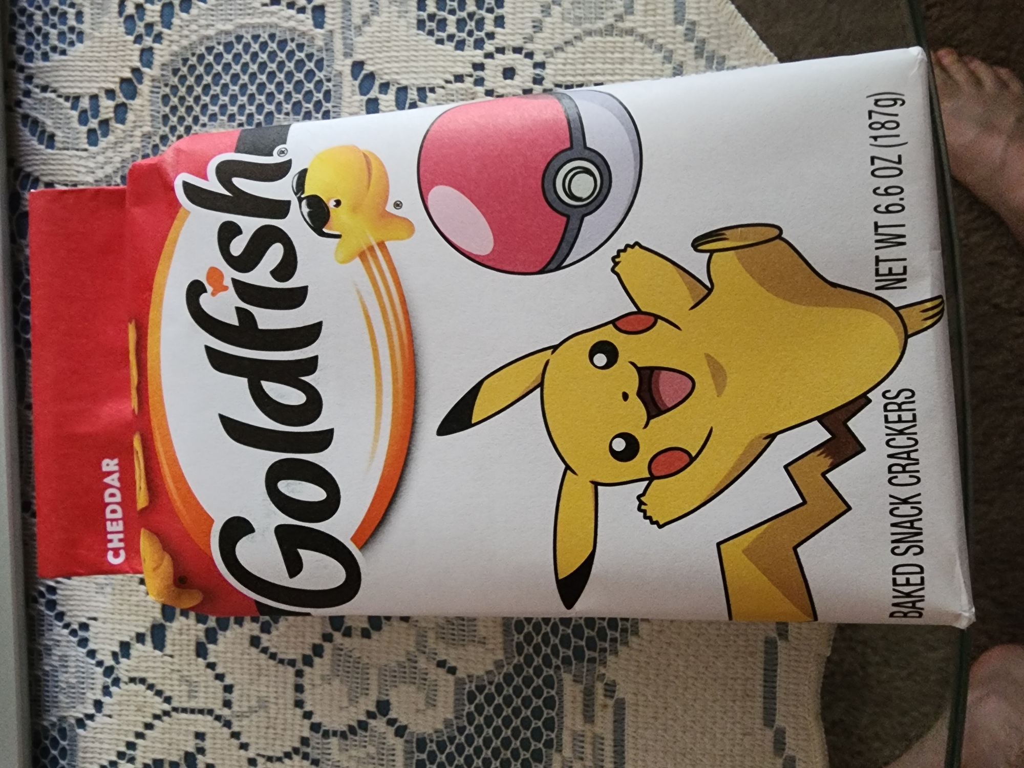 A close-up, eye-level shot captures a bag of Goldfish Cheddar baked snack crackers, featuring a Pokemon collaboration. The bag, predominantly white with a red top flap, is adorned with a large illustration of Pikachu on the front. Pikachu, depicted in its classic yellow and red, is shown mid-jump with its arms raised. To the right of Pikachu, a red and white Poké Ball is illustrated. Above Pikachu, a smaller, cartoonish Goldfish cracker with sunglasses is shown in motion, trailing orange lines. The words "Goldfish" are prominently displayed in large, black lettering across the top of the bag, with a small orange goldfish icon above the letter 'f'. The text "CHEDDAR" is visible on the red flap at the top. Below the main illustration, the text "BAKED SNACK CRACKERS" and "NET WT 6.6 OZ (187g)" are printed in black. The bag rests on a surface with a textured blue and white patterned cloth. The lower right corner of the image shows a partial view of a bare foot. The setting is indoors, with the background suggesting a home environment. The image was taken in Lower Paxton Township, United States of America.