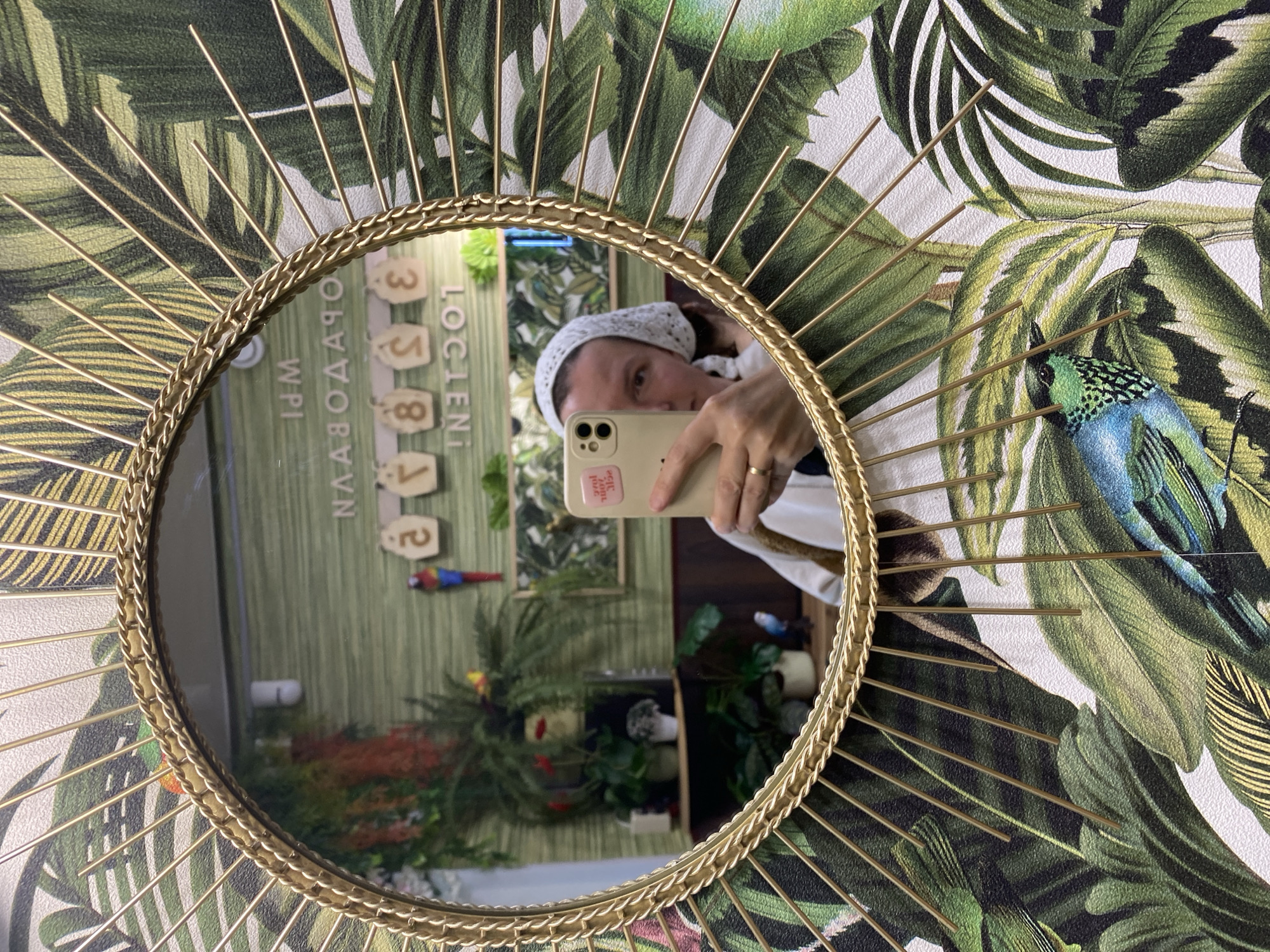 A selfie is taken in a round mirror with gold radial spikes against a tropical-themed wallpaper. The reflection in the mirror shows an interior setting. A sign is visible, showing the city name "Chaikovsky" in Russian: "Чайковский" and local time, "WPI 1PM".