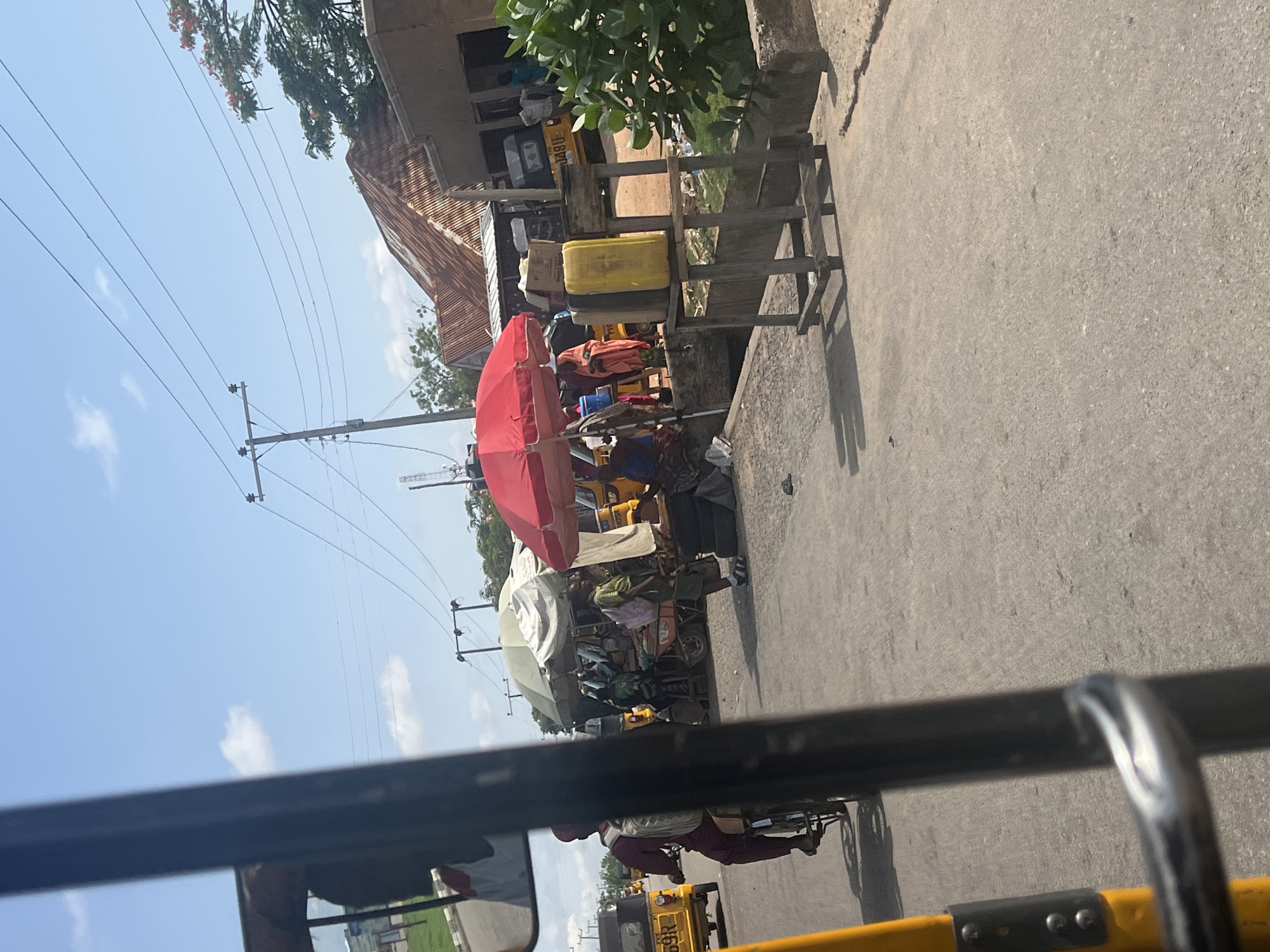 This image depicts a street scene in Jalingo, Nigeria. The view is from inside a yellow vehicle, likely a rickshaw, with a metal bar in the foreground and the yellow vehicle's interior visible on the left. The street is paved with asphalt and shows signs of wear.
In the background, a bustling market area is visible. Several yellow rickshaws, a common mode of transport in the region, are parked or moving. People are seen interacting at market stalls set up under umbrellas. One stall features a prominent red umbrella. Items like crates, containers, and bundles are displayed for sale. A person is riding a motorcycle wearing a maroon outfit and a backpack. Another person in a green shirt and dark pants is walking with a backpack. Several yellow jerrycans are stacked on a wooden stand. In the distance, buildings with corrugated iron roofs and overhead power lines are present, suggesting an urban or semi-urban environment. The sky is blue with some scattered white clouds. The overall impression is of a lively market day in Jalingo.
