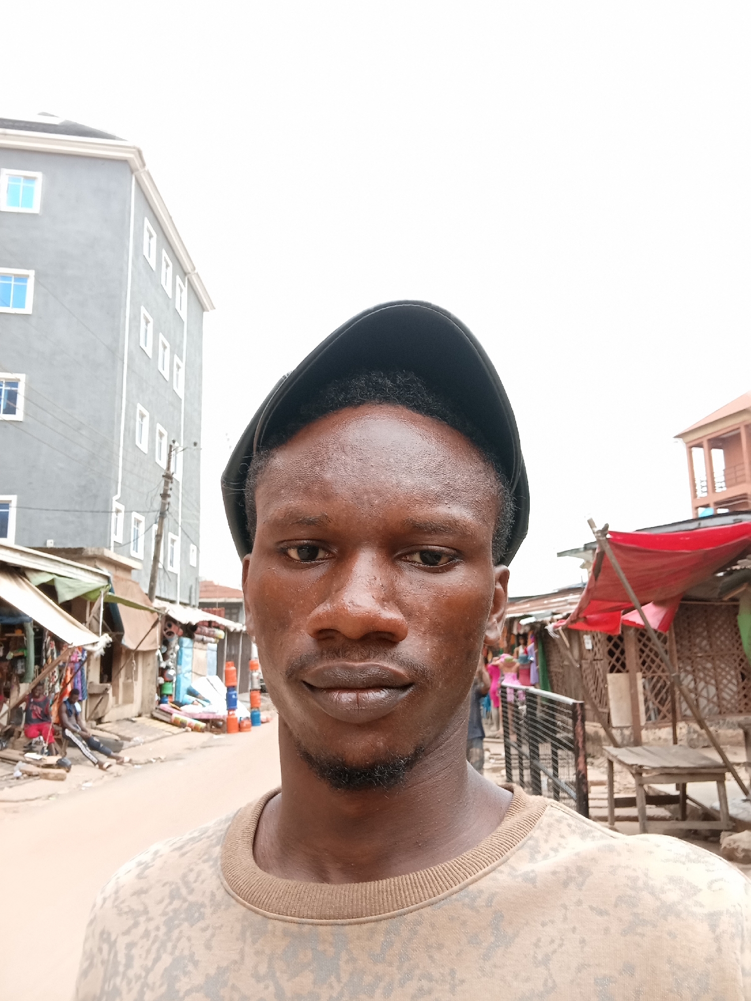 A man is captured in a close-up selfie, standing on a bustling street in Onitsha, Nigeria. He is positioned centrally, gazing directly into the camera with a neutral expression. He has dark skin, a short beard or stubble, and wears a dark cap, possibly backward, and a light-colored sweatshirt featuring a subtle camouflage-like pattern. His forehead and cheeks appear slightly shiny, possibly from sweat or humidity.
The background reveals a vibrant, somewhat chaotic street scene under an overcast sky. To the man's left, a multi-story grey building with white window frames, some holding blue-tinted glass, dominates the skyline. Below it, several market stalls line the unpaved road, constructed from simple materials with corrugated iron roofs. A utility pole with wires is also visible.
On the right side of the street, more market stalls with prominent red tarpaulin awnings and wooden structures can be seen. Various goods, though indistinct, are displayed in the stalls. Blurred figures of other people are visible further down the street, indicating ongoing activity and pedestrian traffic. The scene depicts a lively urban market environment during the daytime, characterized by diffused natural light. No discernible text is visible within the image.